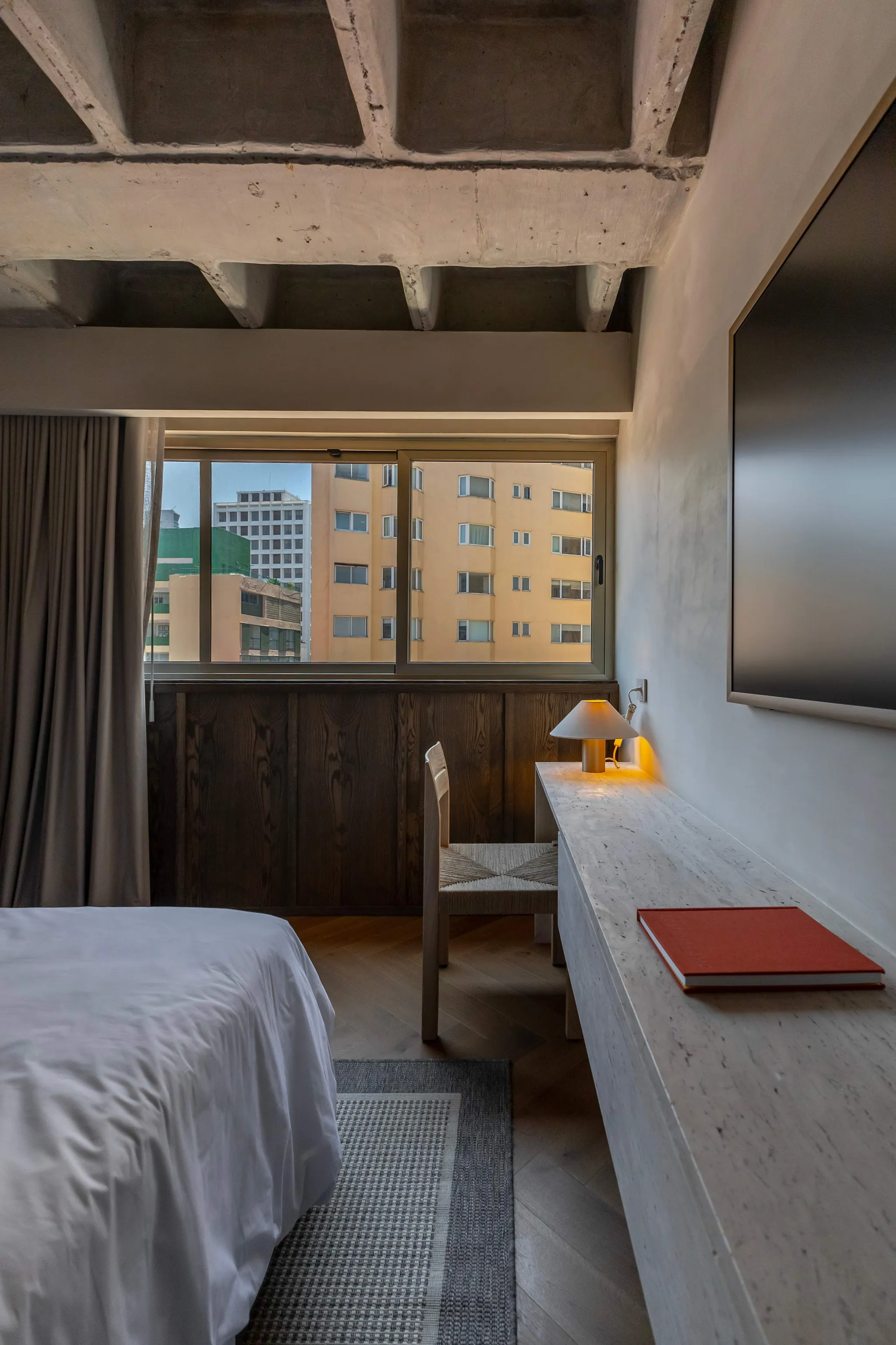 A bedroom with a bed, desk, chair, and window view of city buildings.