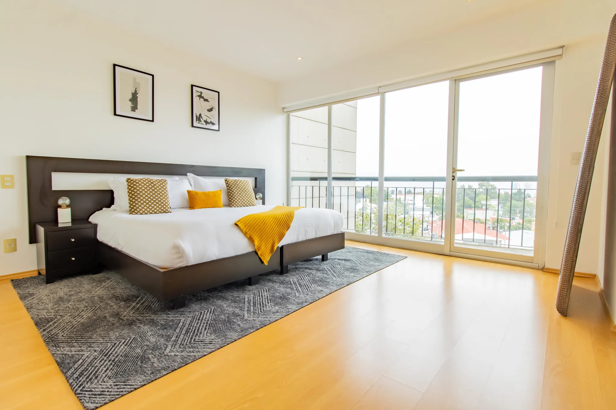 A modern bedroom features a bed with white linens and accent pillows, a nightstand, and a large wind
