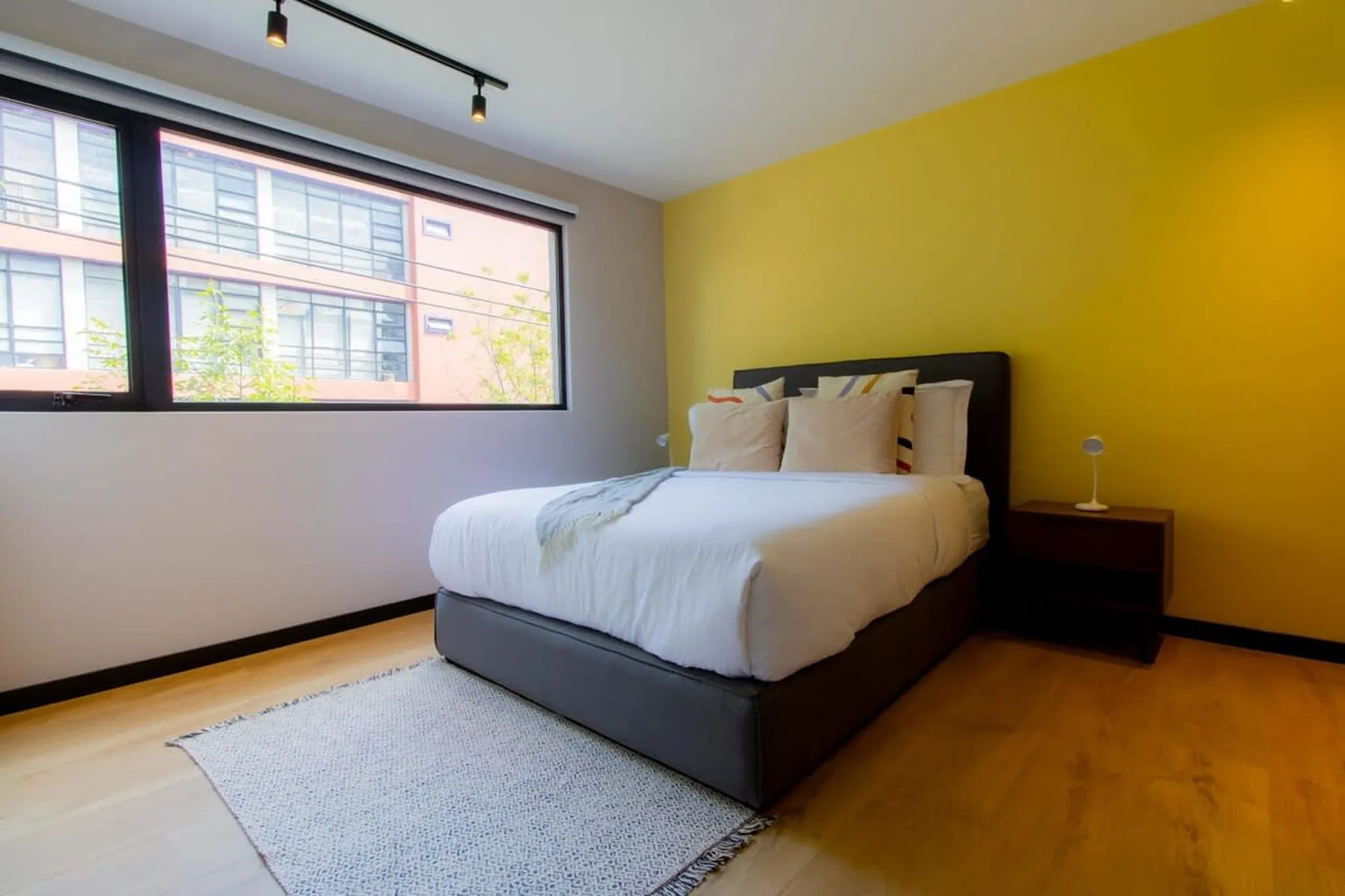 Modern bedroom with a queen-size bed, large window with city view, and yellow accent wall.
