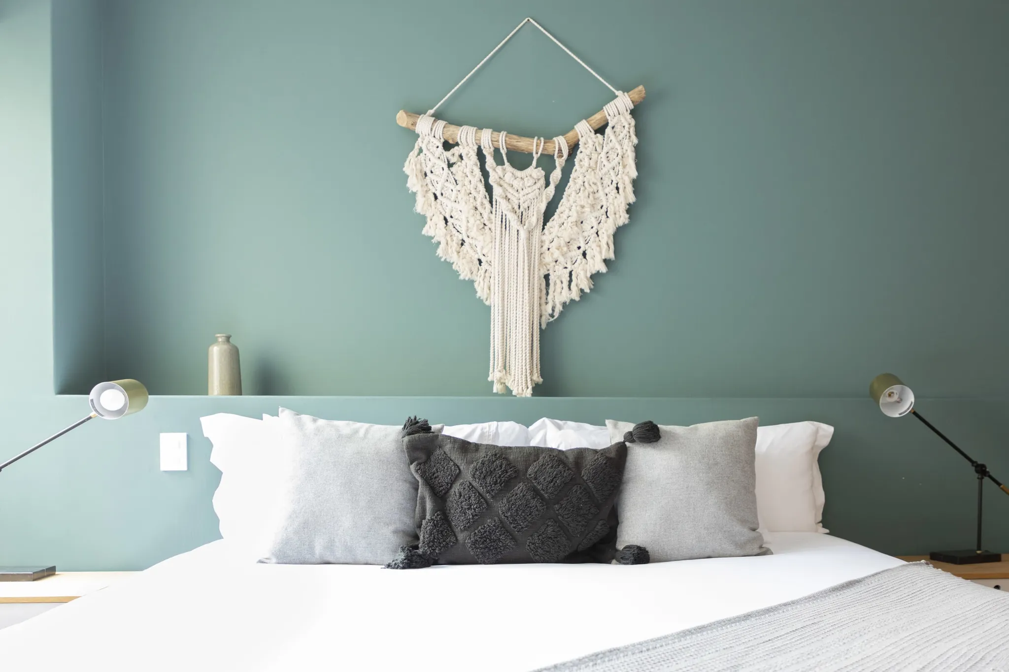 A macrame wall hanging above a bed with grey pillows and a grey throw.