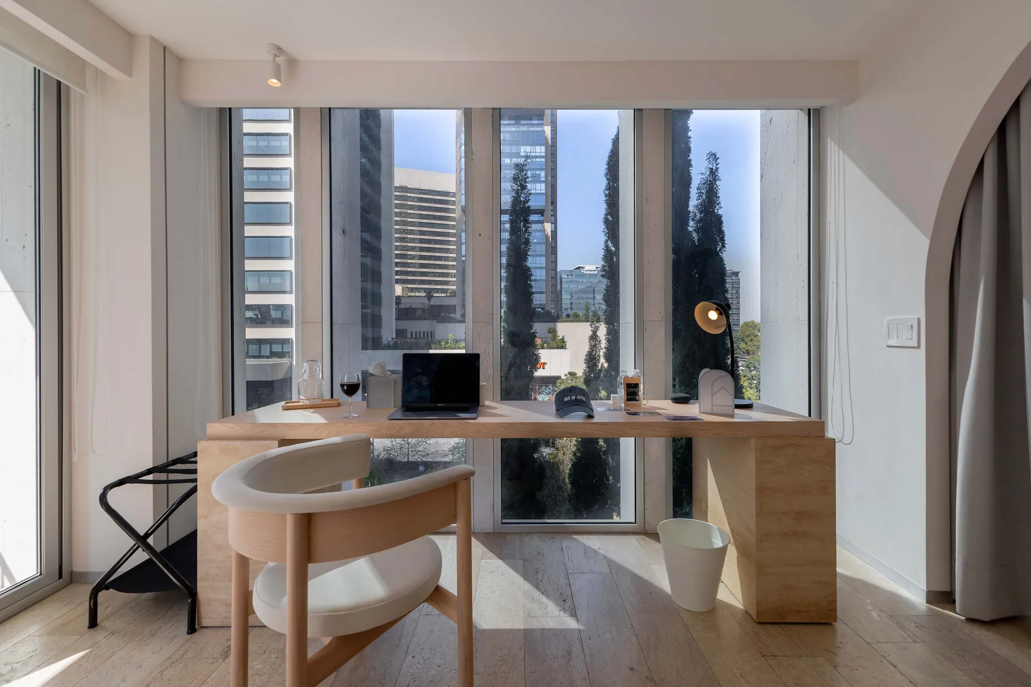 A modern office desk with a laptop sits by a large window overlooking a city skyline.