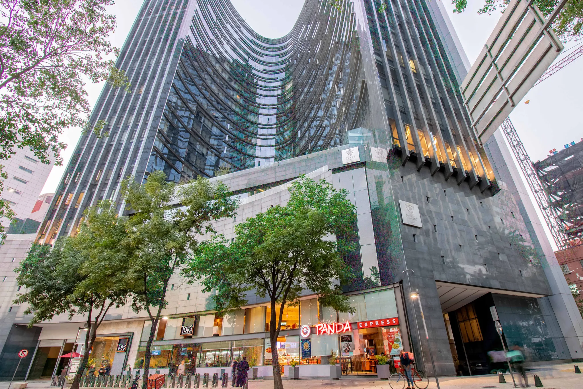 A modern, curved glass building with trees and storefronts at its base.