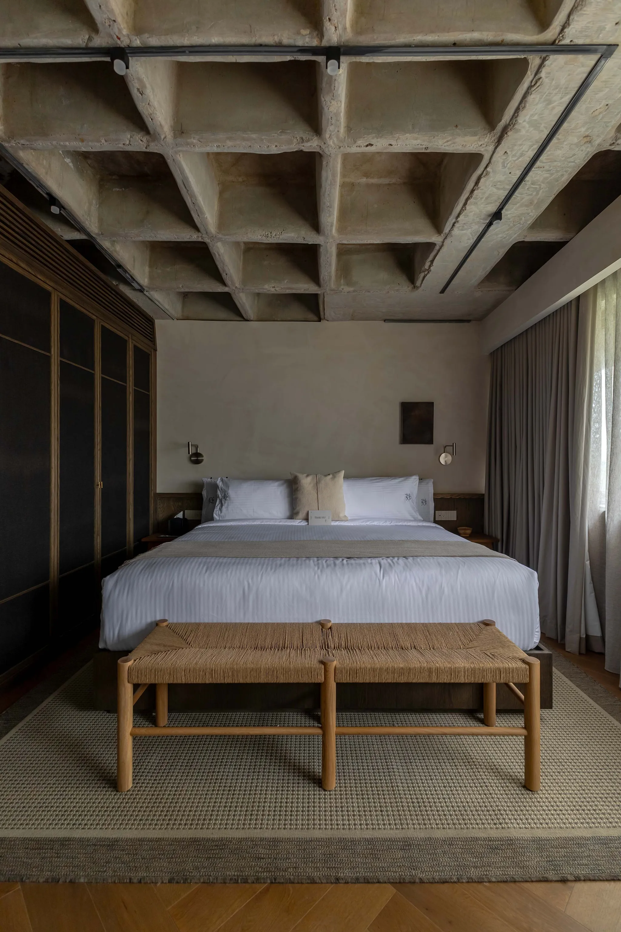 A modern bedroom with a concrete waffle ceiling, a large bed, and a woven bench.