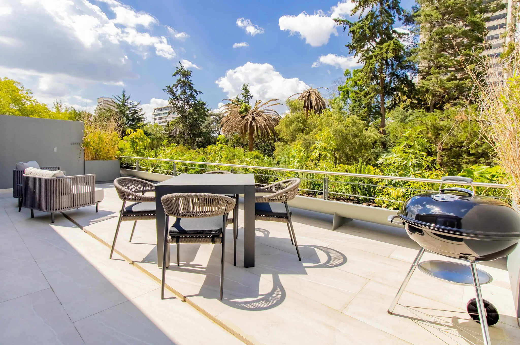 A patio with a table, chairs, and a grill, surrounded by trees and a building.