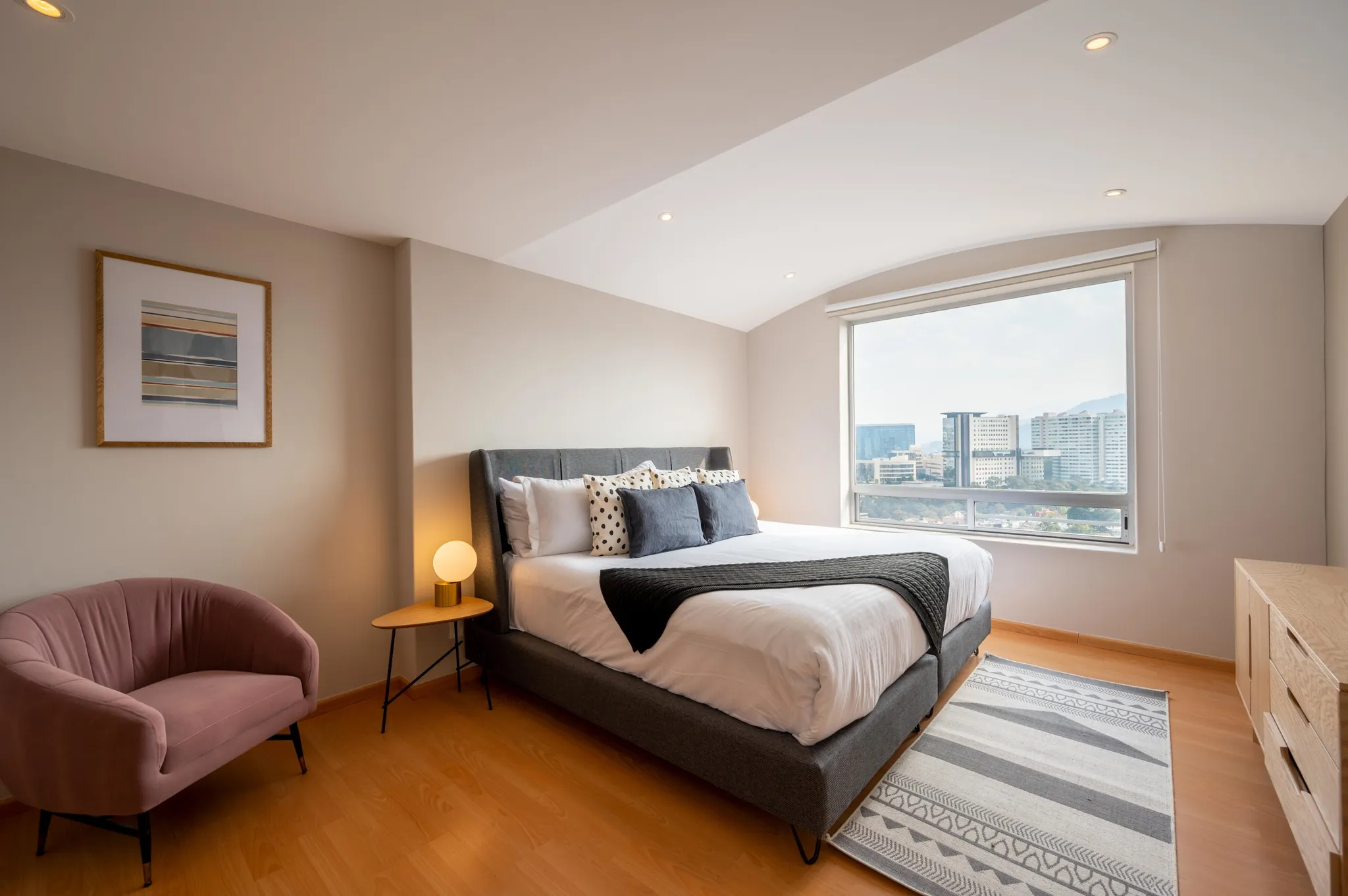 A bedroom with a bed, a pink chair, and a large window overlooking a city.