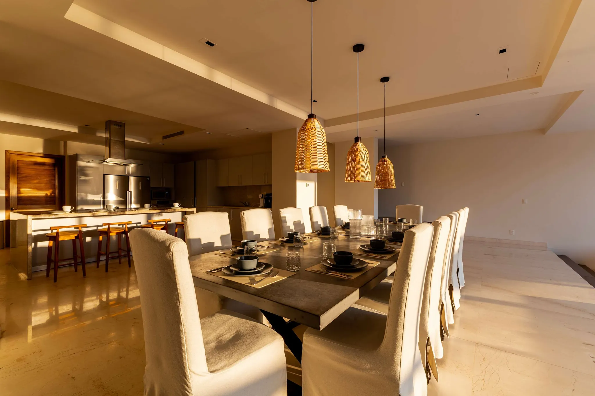 A dining room with a long table set for a meal, with a kitchen in the background.