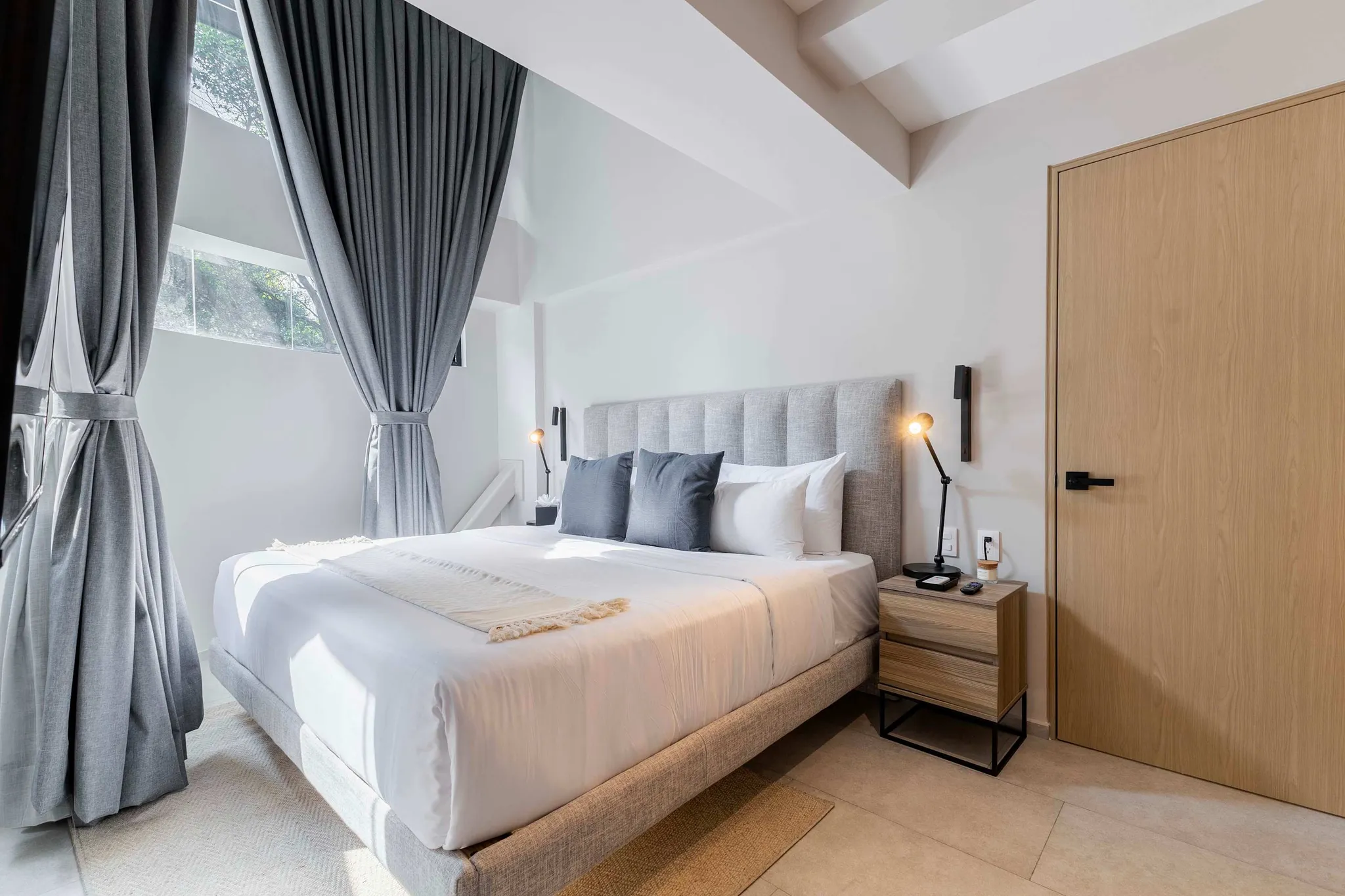 A modern bedroom with a large bed, grey curtains, and a wooden door.