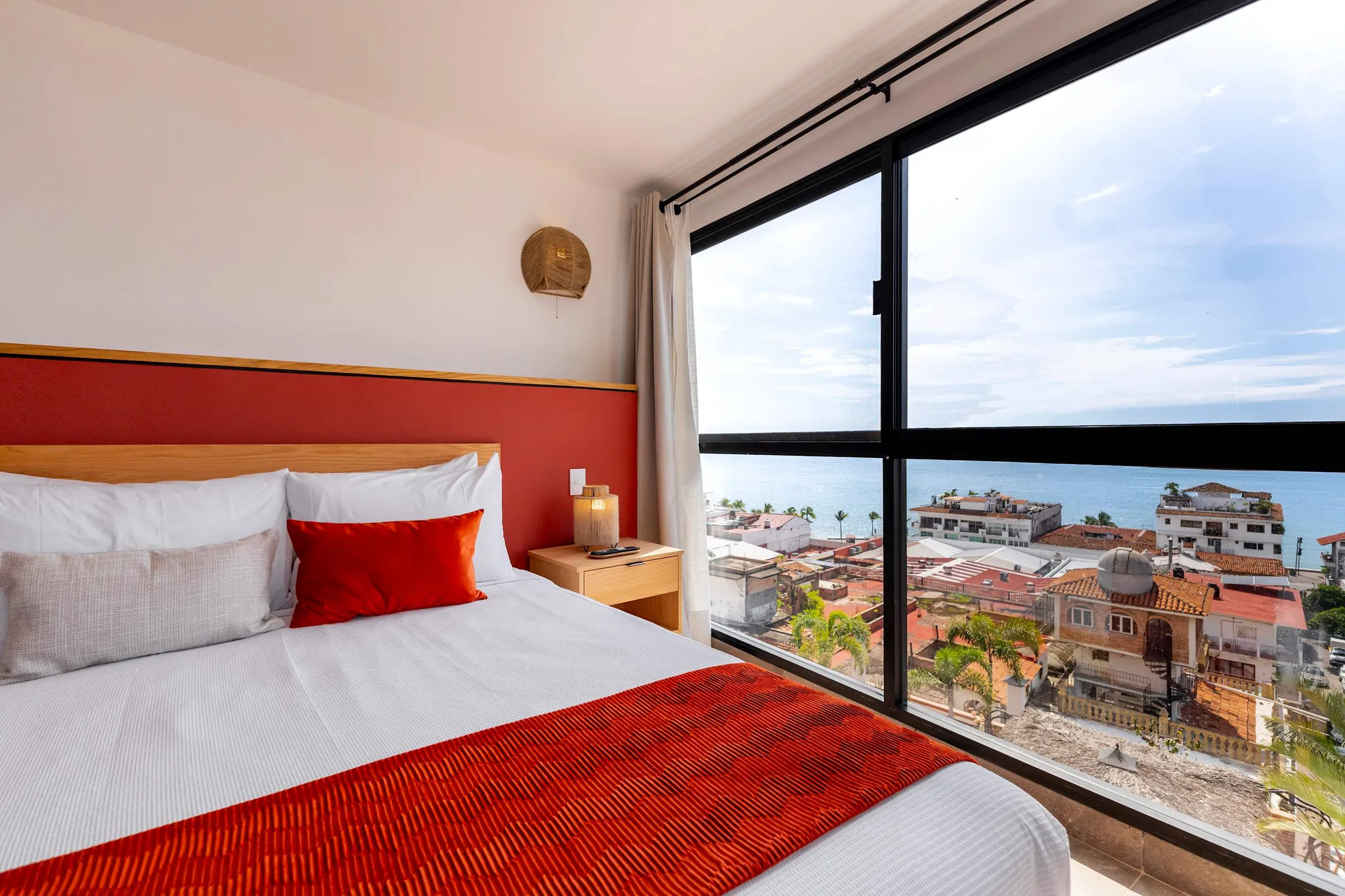 A bed with white linens and a red throw, next to a large window with a city and ocean view.
