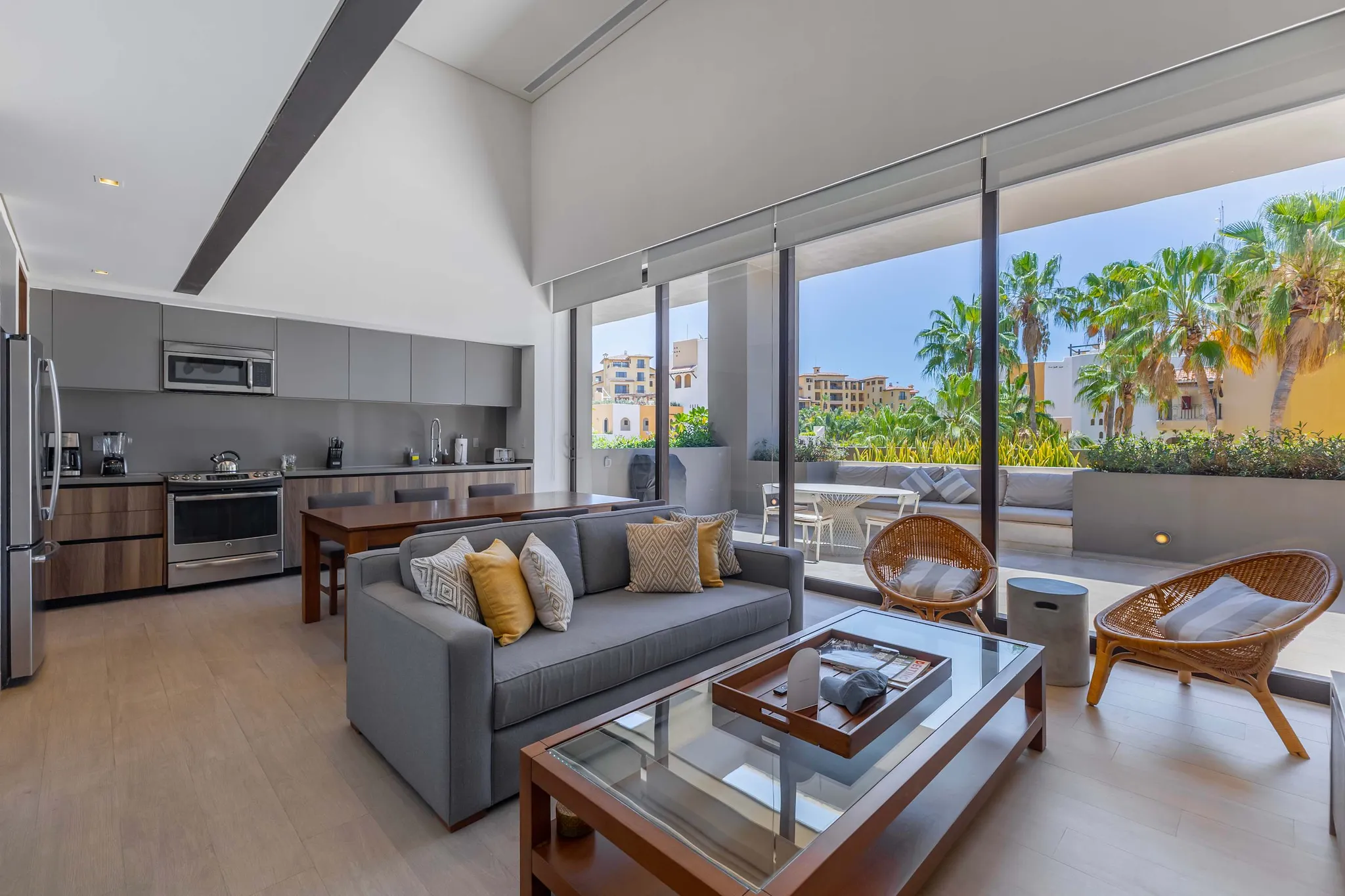 Modern living room with gray sofa, coffee table, and kitchen area, opening to a balcony with palm tr