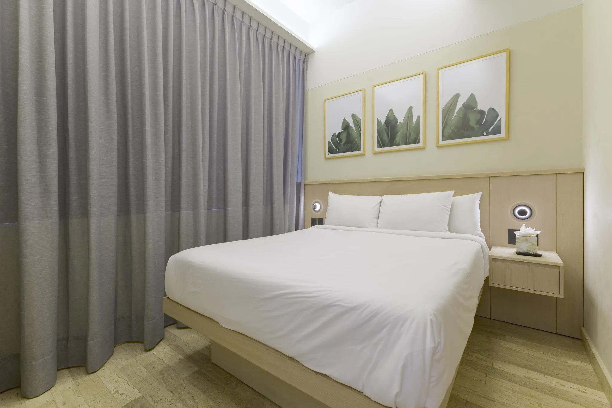A modern bedroom with a queen bed, grey curtains, and tropical leaf artwork.