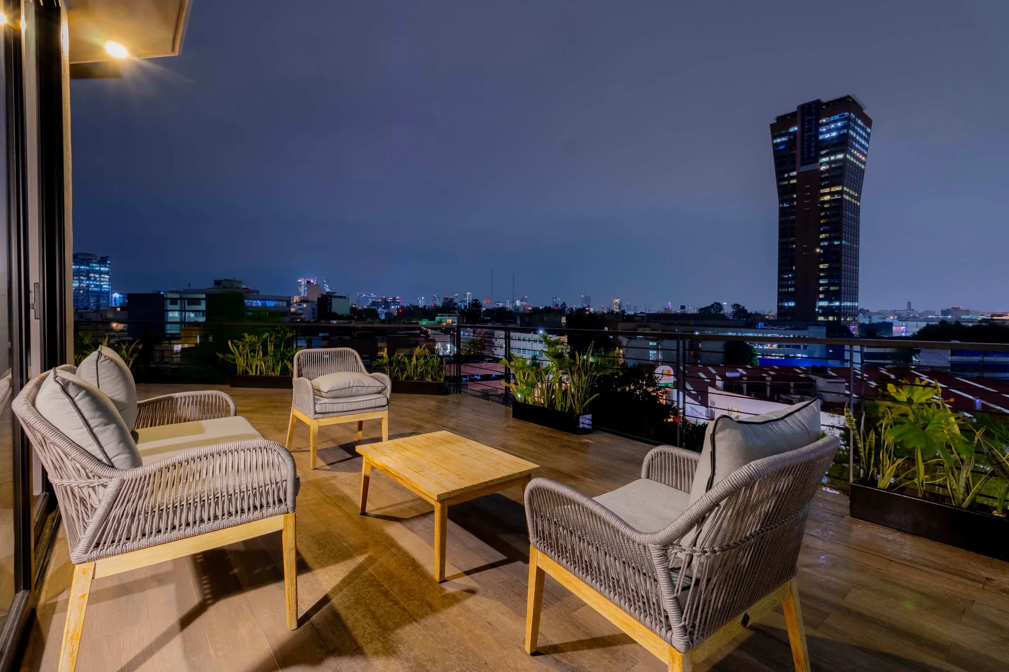 A nighttime rooftop patio with wicker furniture overlooks a city skyline.