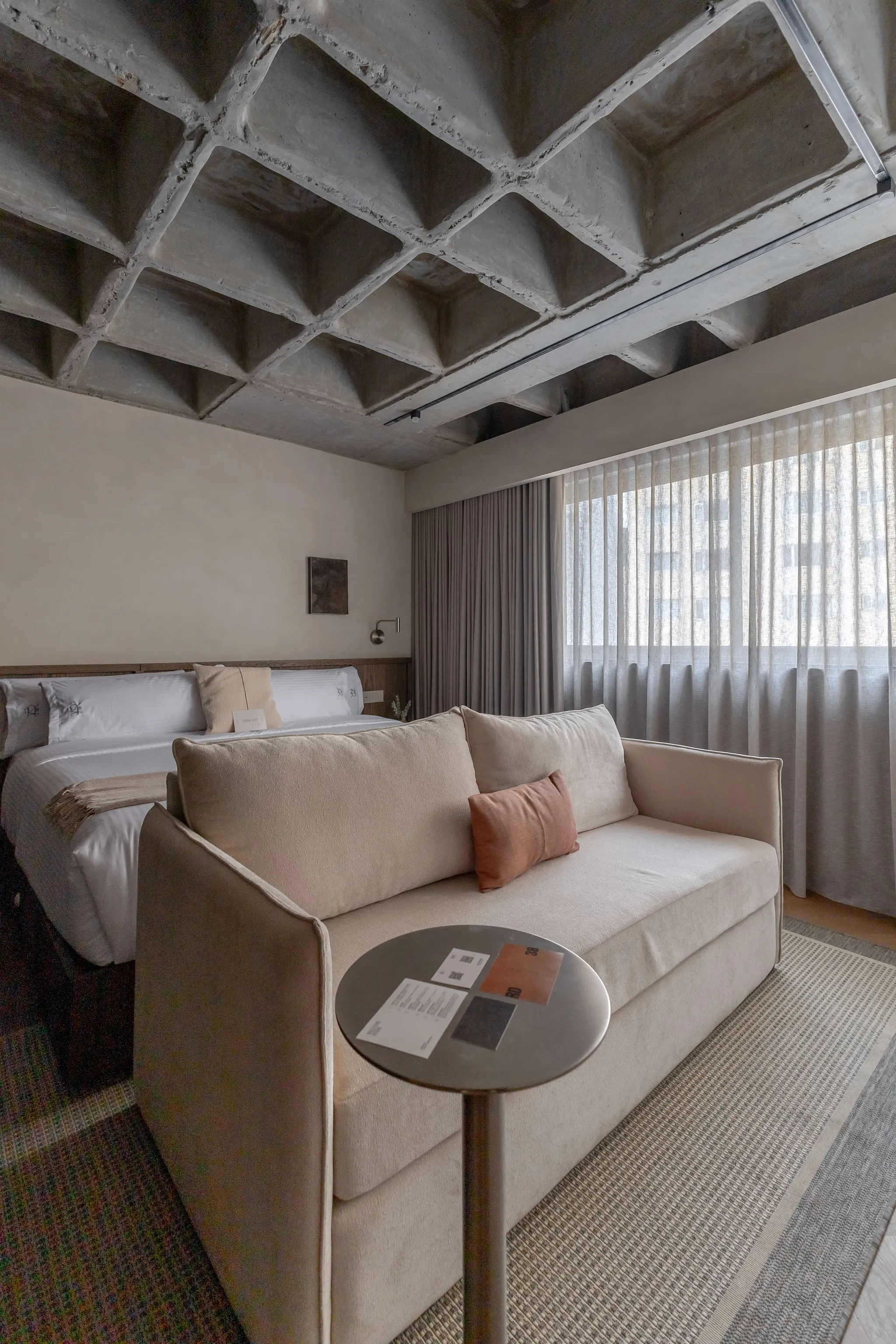 A modern bedroom with a beige couch, a bed, and a textured concrete ceiling.