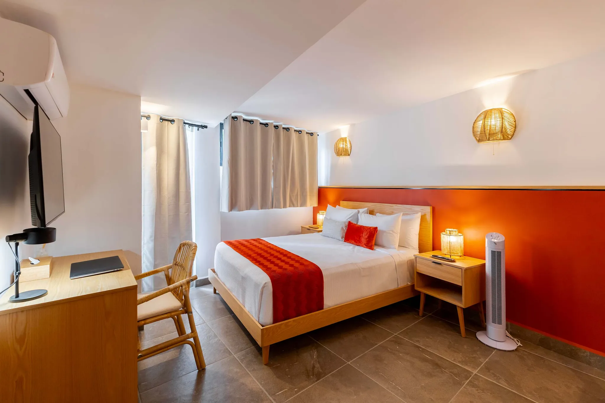 A well-lit hotel room with a bed, desk, chair, and fan.