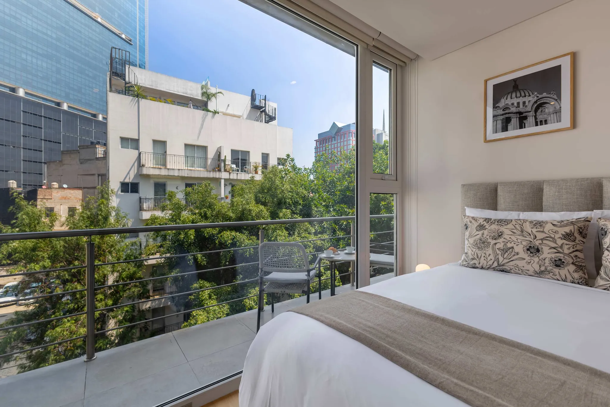 Modern bedroom with city view from balcony, featuring a bed, pillows, and a table with coffee.