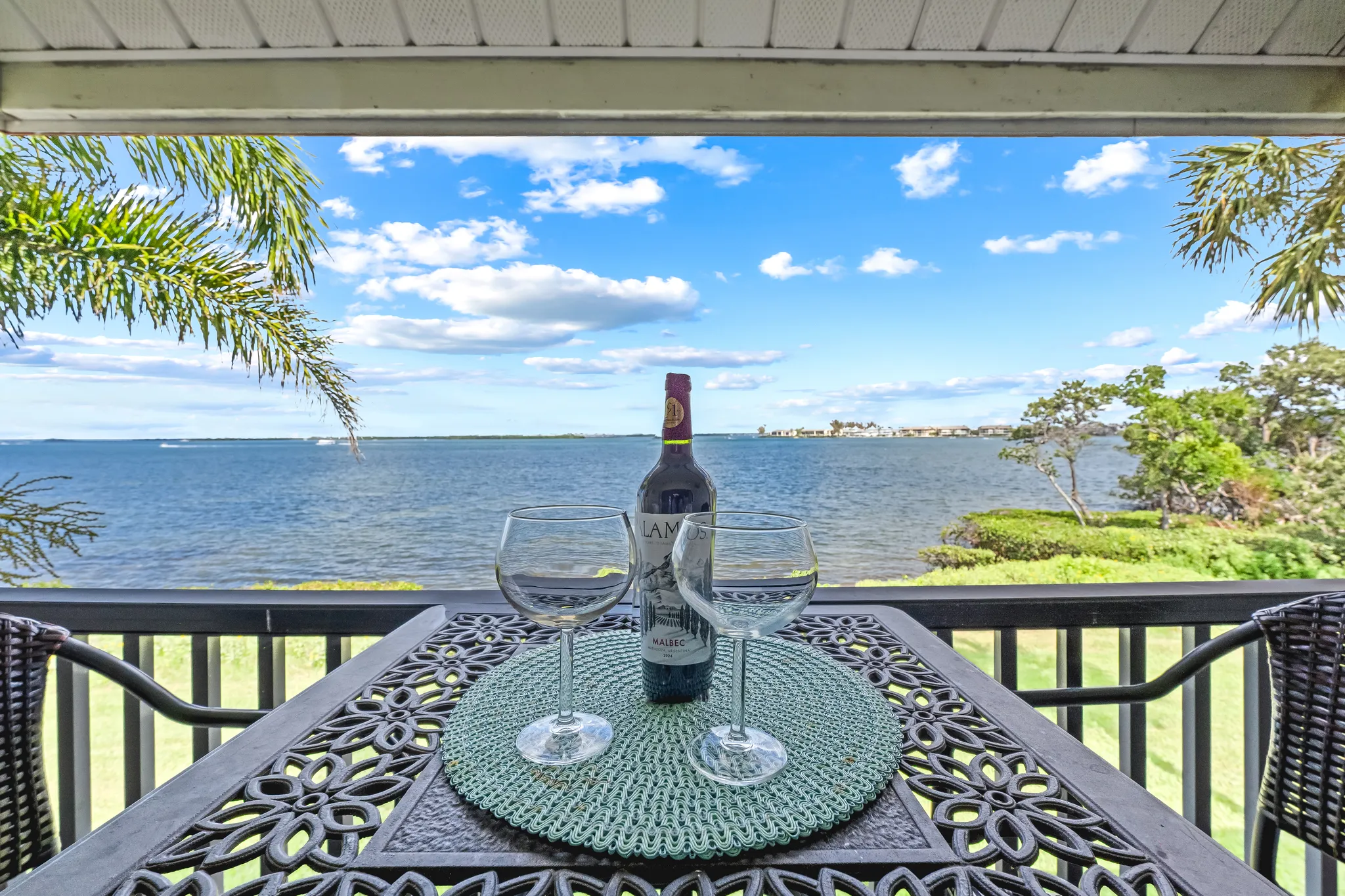 Covered patio with wooden bistro set, wine bottle and lantern centerpiece, overlooking a grassy lawn and tranquil bay view