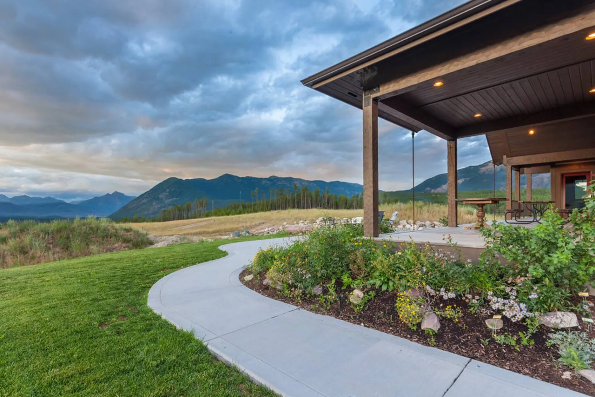 A modern house with a porch overlooks a vast mountain landscape at dusk.