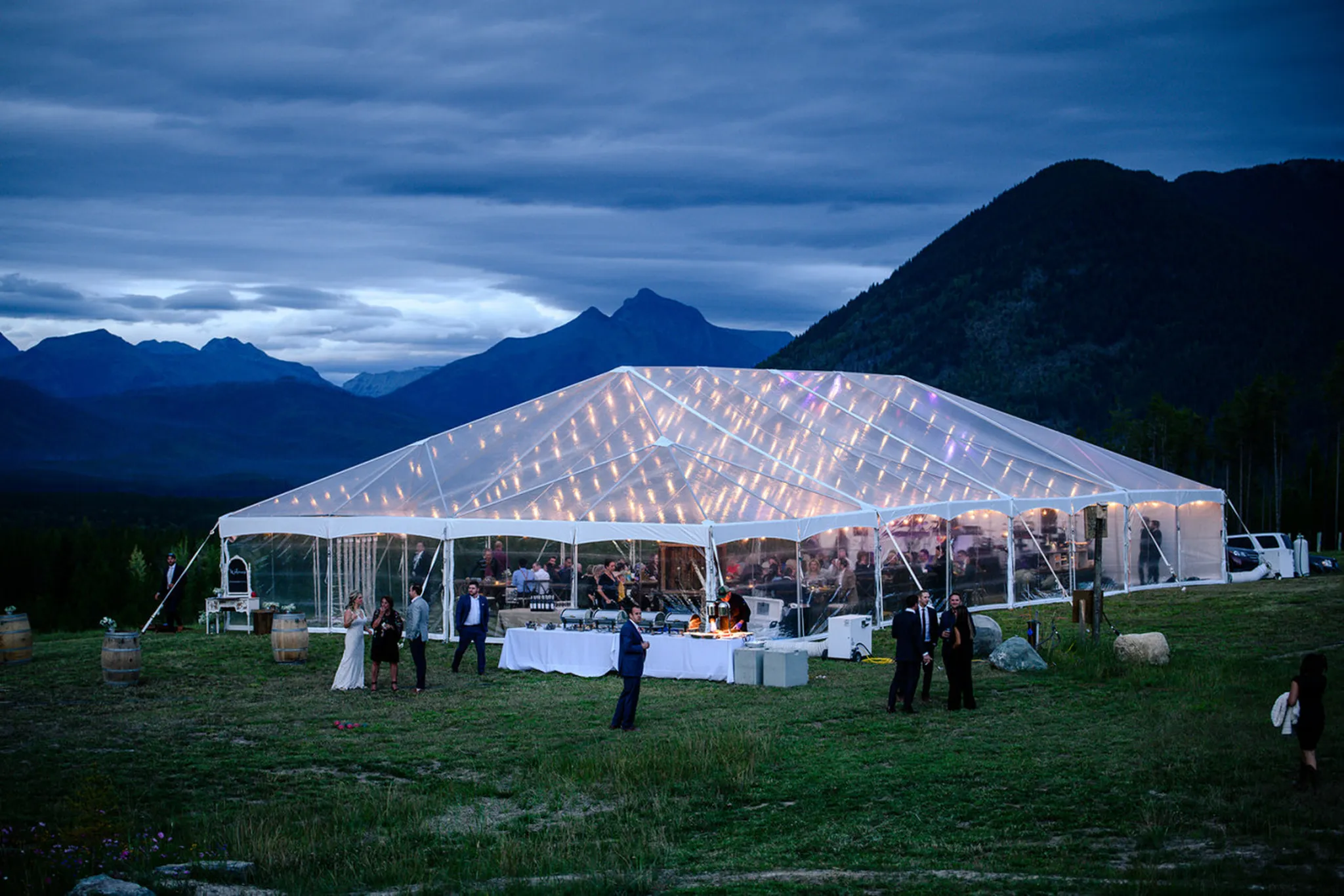 An outdoor wedding reception under a clear tent with lights at dusk, mountains in the background.
