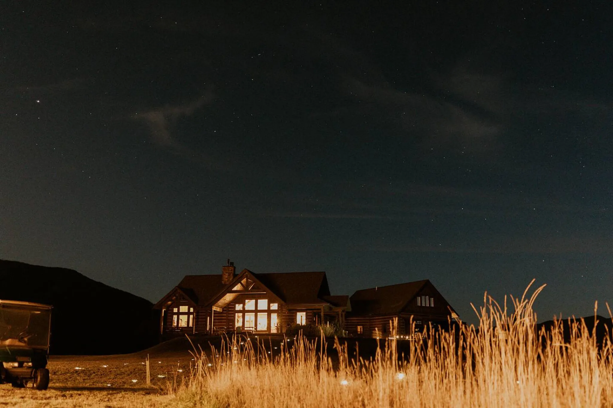A cozy log cabin glows warmly under a starry night sky, with tall dry grass in the foreground.