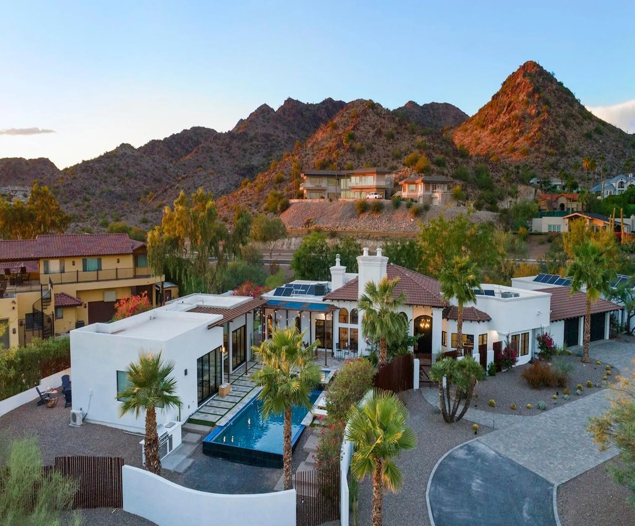 Aerial view of a desert hillside estate with pool, palm trees and tiled-roof villa backed by rugged sunlit mountains at dusk
