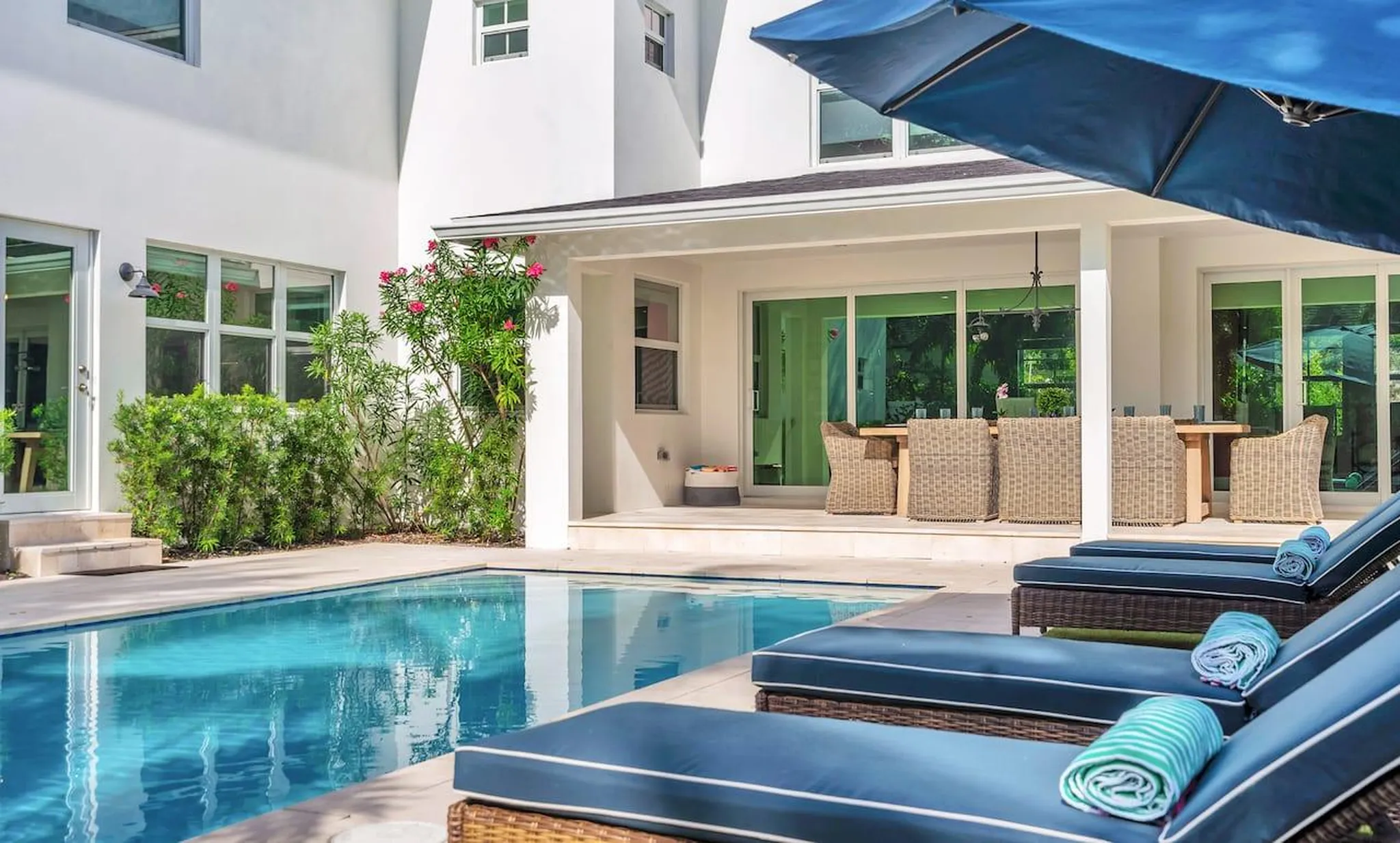 Backyard pool with navy loungers and blue umbrella beside a covered patio with wicker dining set and flowering shrubs