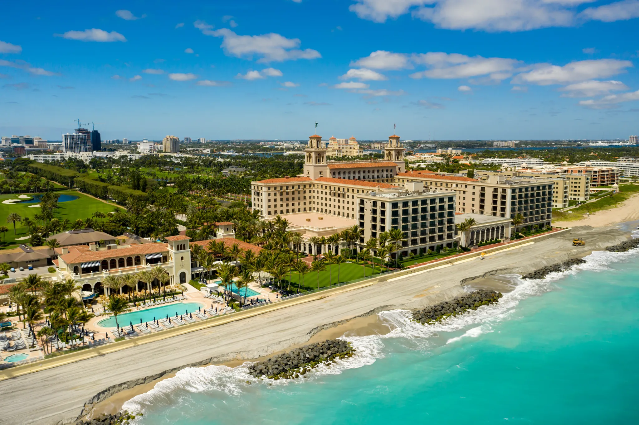Aerial view of a grand oceanfront resort with twin towers, palm-lined pools, and turquoise sea along a rocky shoreline