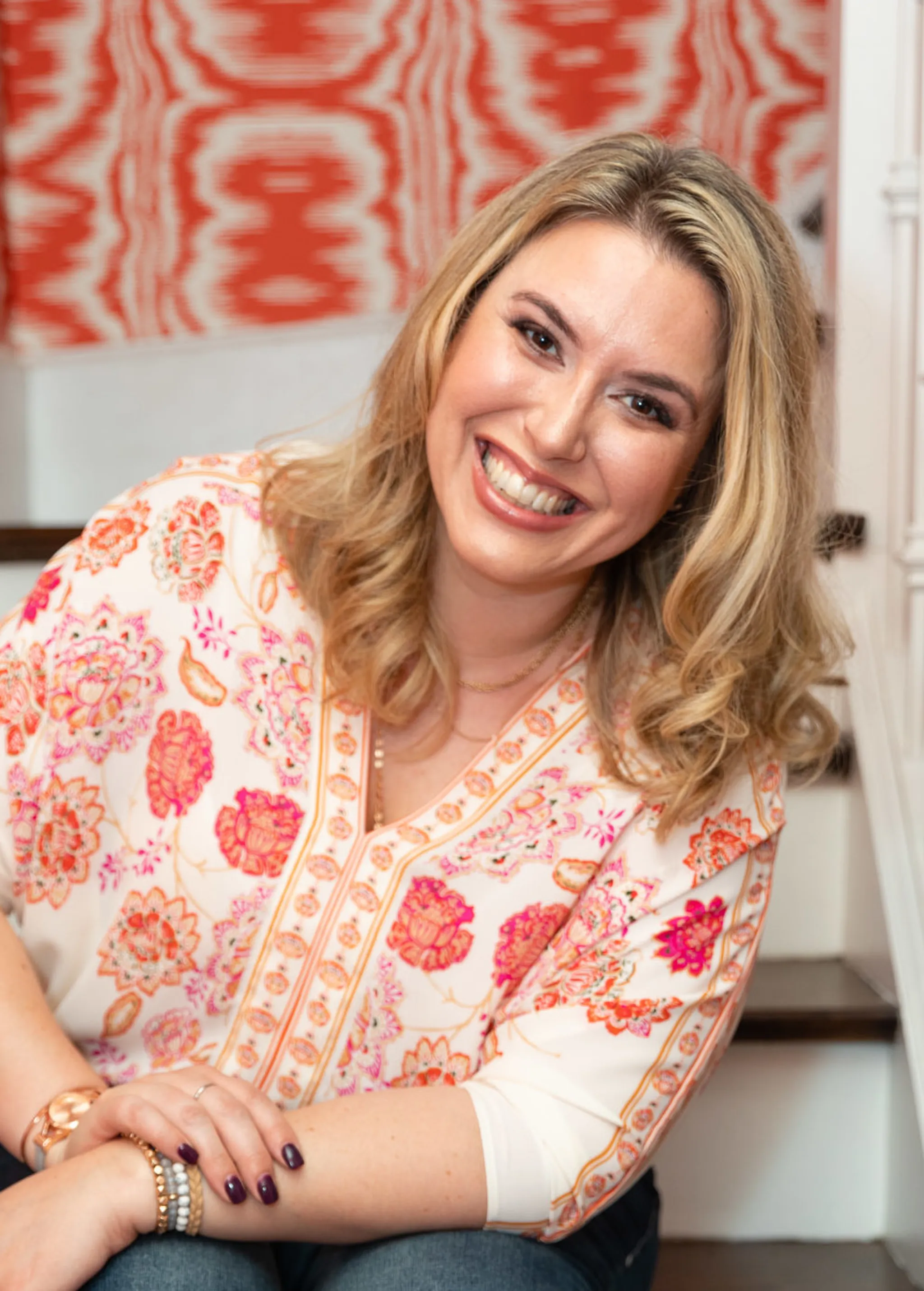 Smiling woman with wavy blonde hair wearing a pink and orange paisley blouse, seated in front of a coral ikat-patterned tapestry