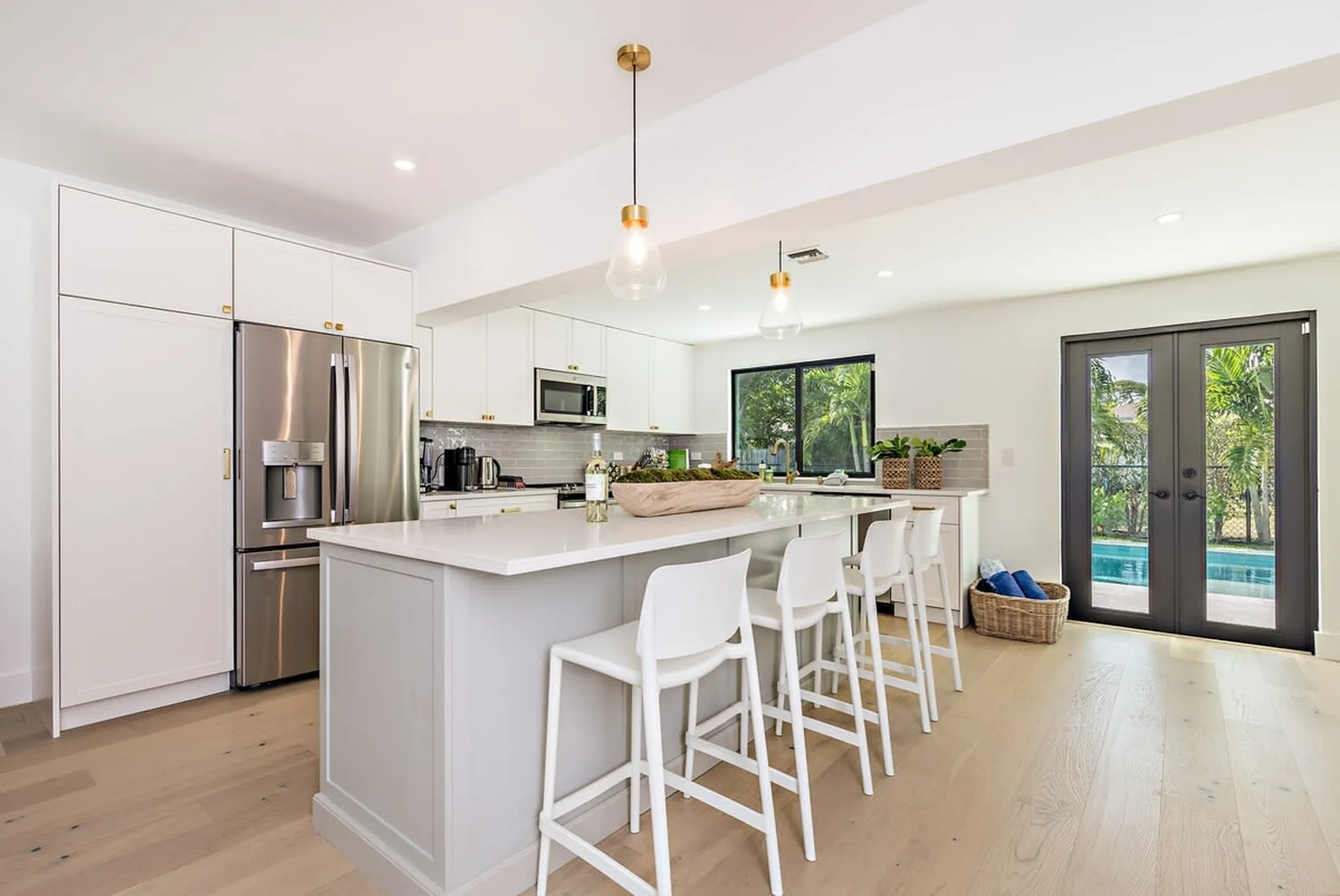 White kitchen with a large island, brass pendant lights, and French doors opening to the pool