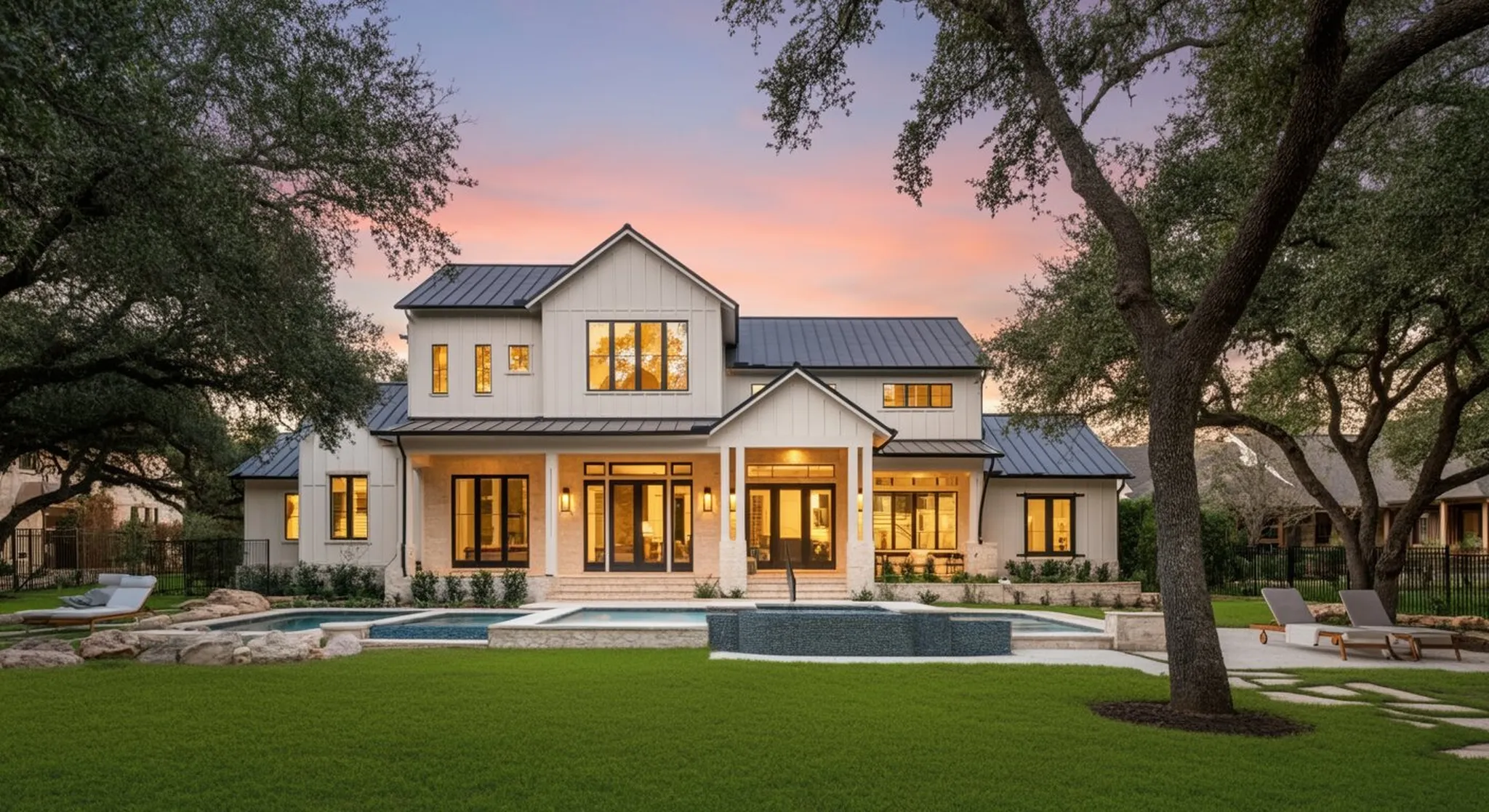 Modern farmhouse exterior at dusk with glowing windows, manicured lawn, pool, and oak trees framing the home under a pink sky