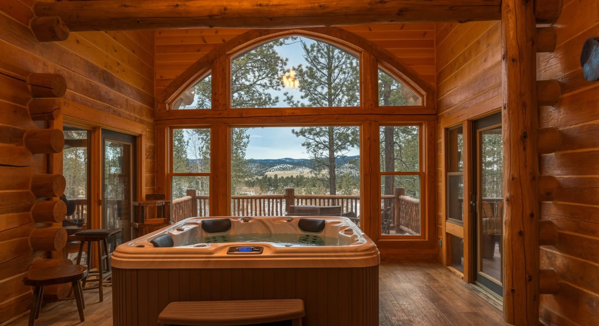 Indoor hot tub in a log cabin sunroom with arched picture windows framing forested mountain views and a wraparound deck