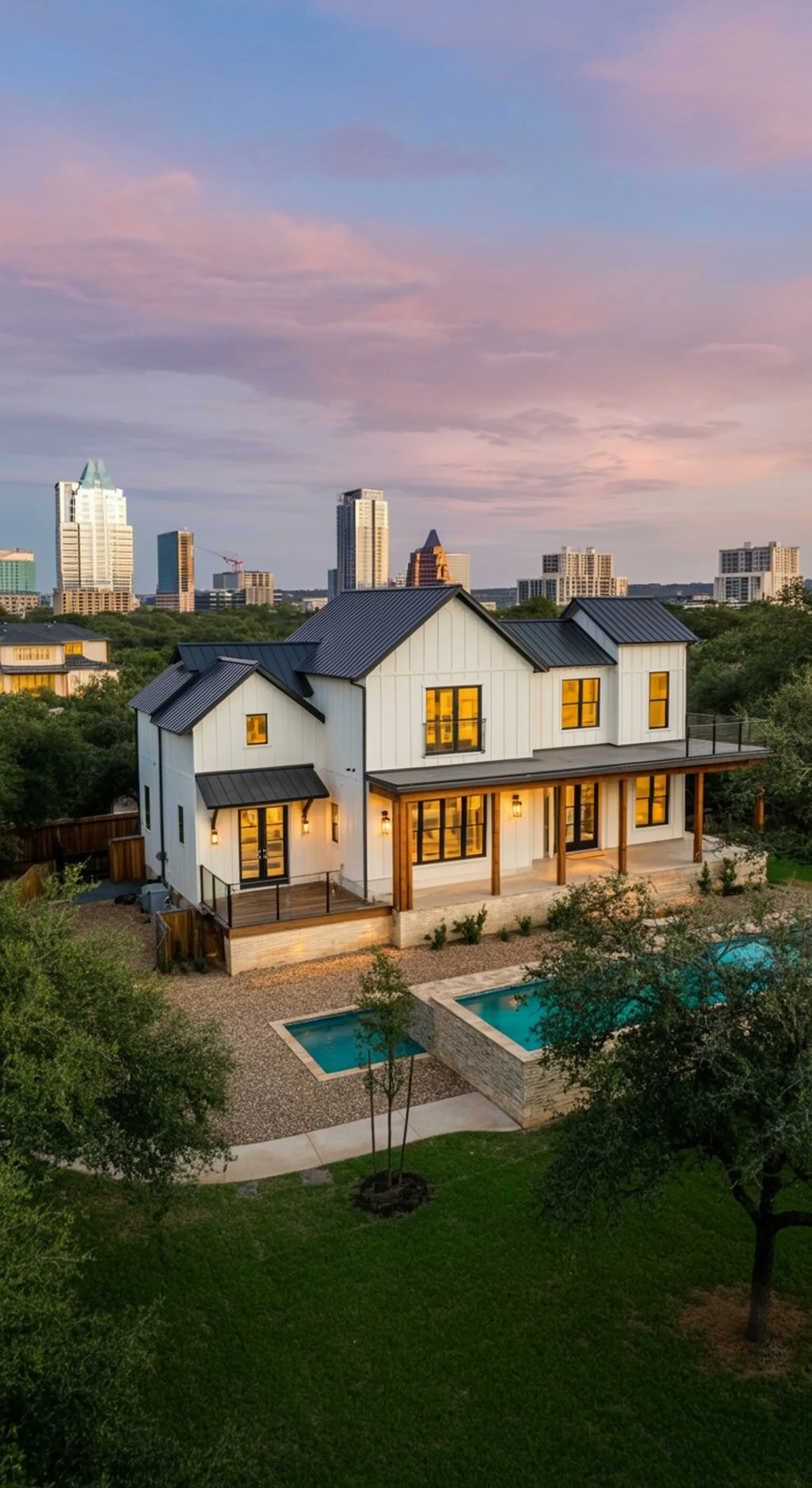 Modern white farmhouse with metal roof and pool at dusk, downtown skyline glowing under pink sunset clouds in the distance