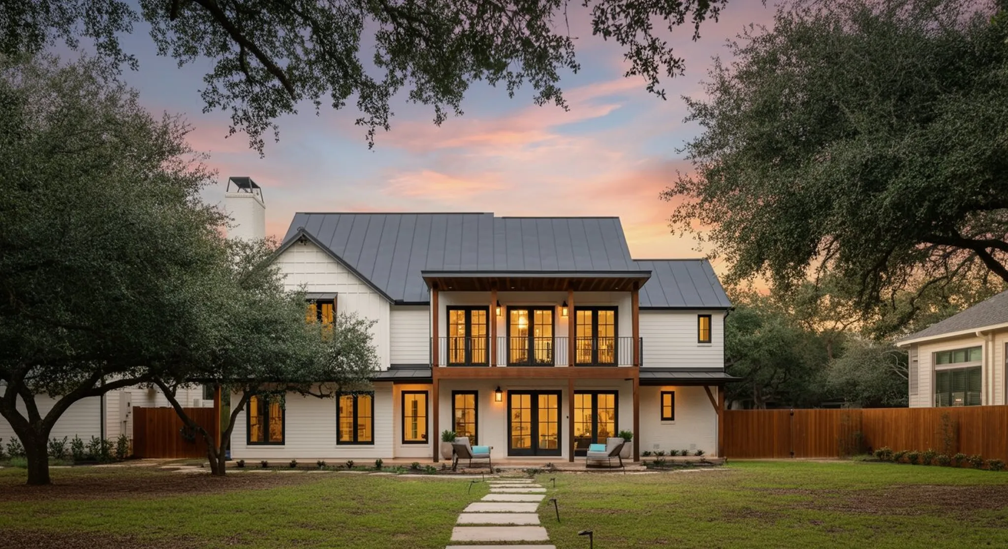 Two-story modern farmhouse at dusk with metal roof, glowing windows, upper balcony, and stone path through a manicured lawn