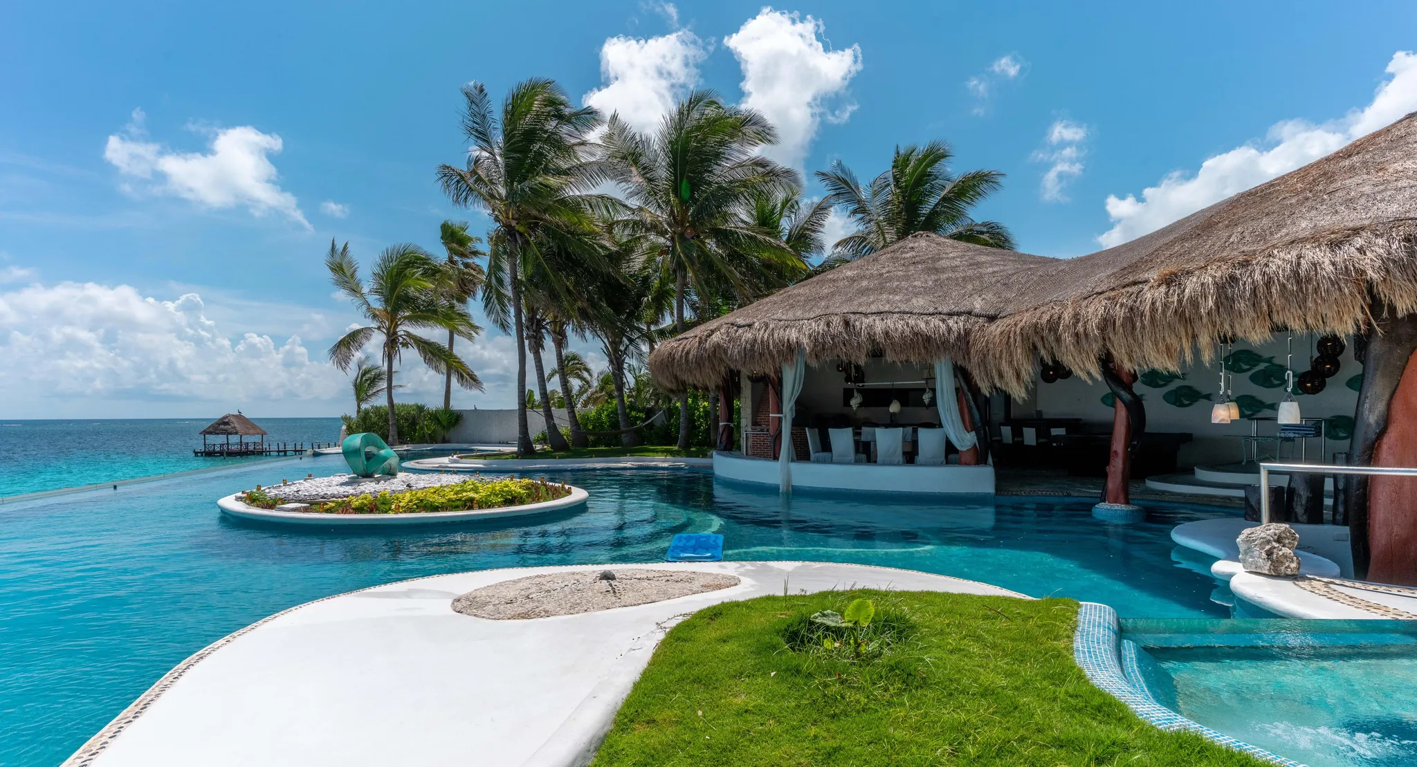 Thatched-roof oceanfront villa with curved infinity pool, palm trees, and green lawn under a blue sky by the sea