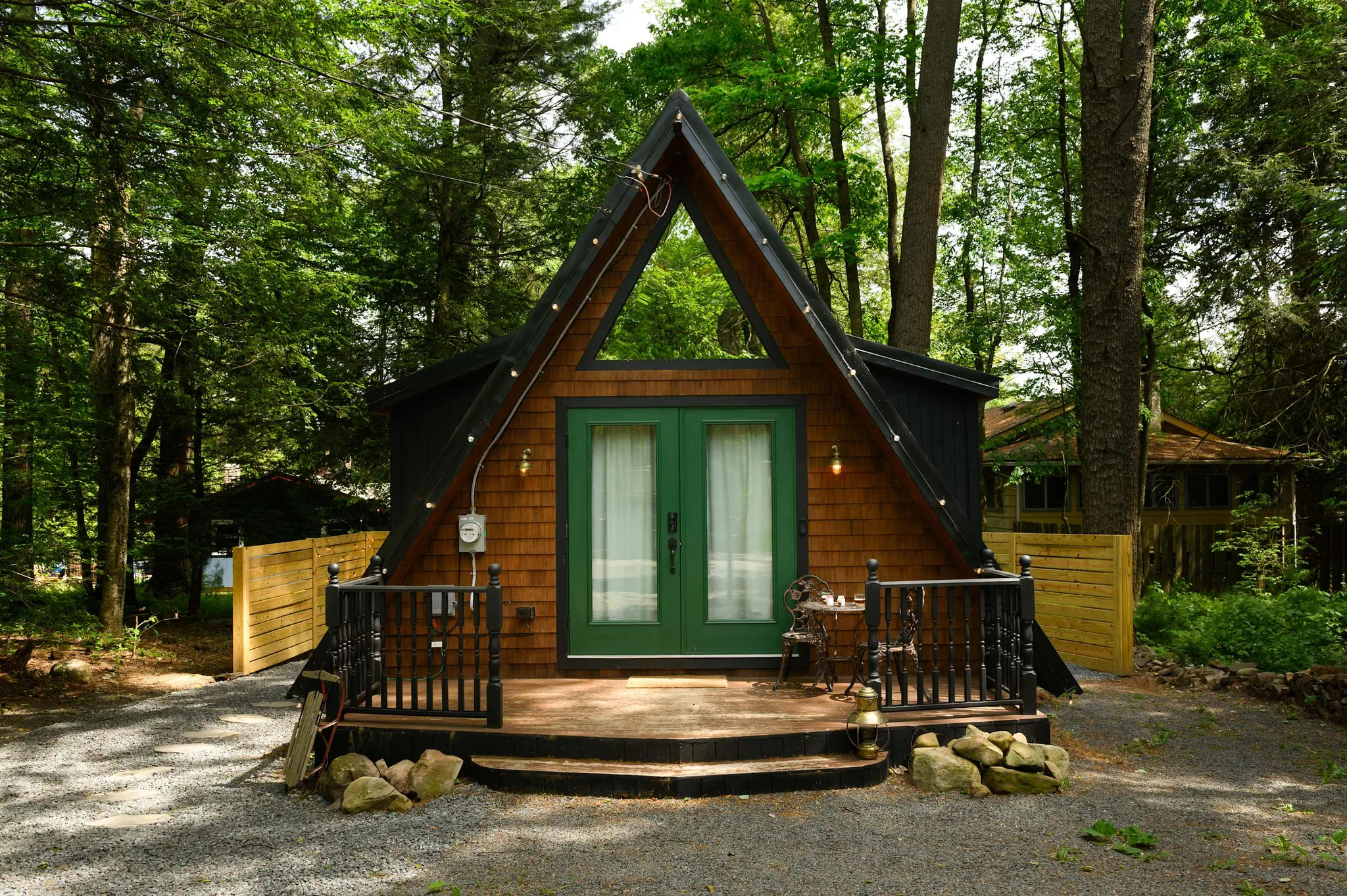 A-frame wooden cabin with green double doors and string lights nestled among tall trees with a gravel path leading to the porch