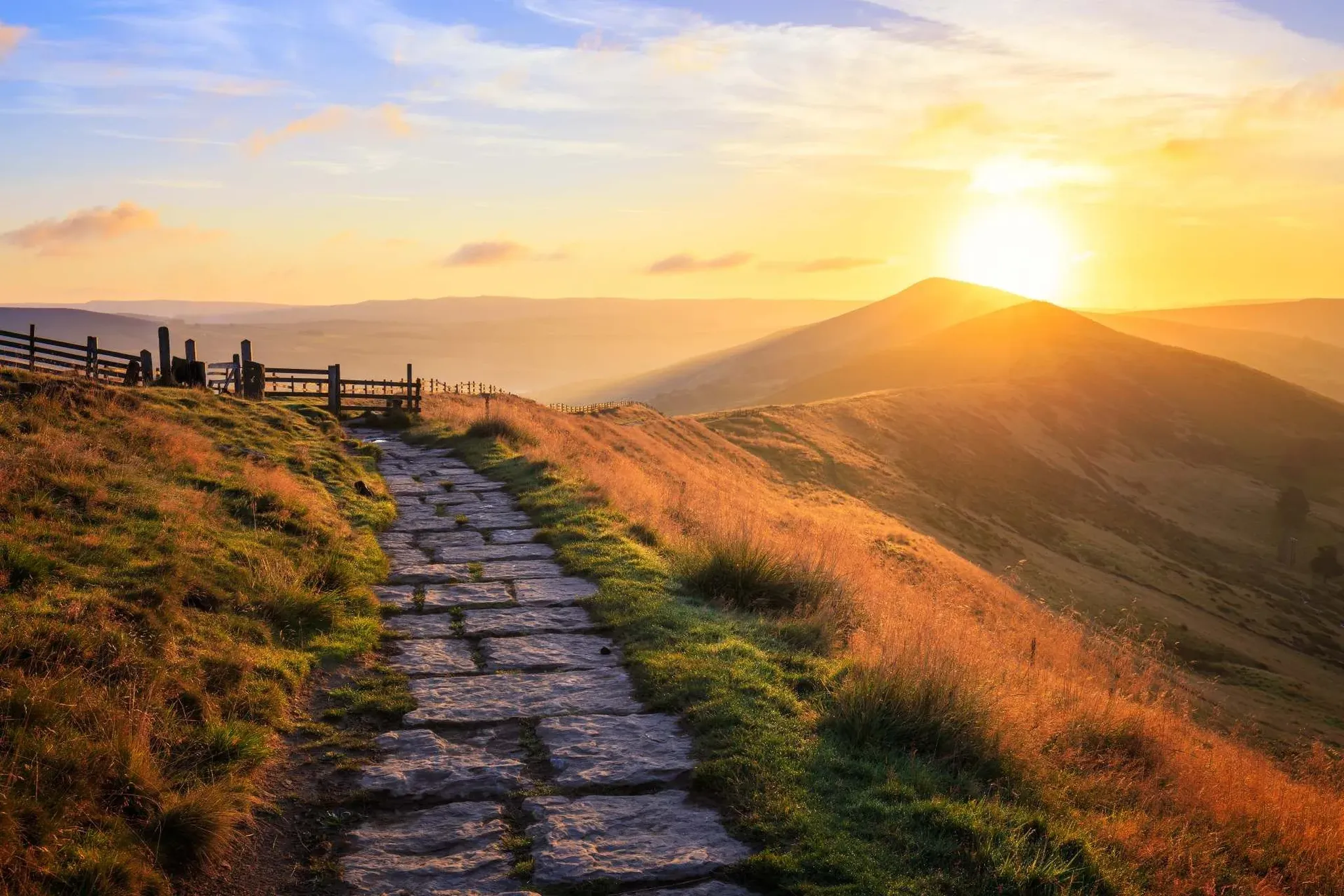 Stone footpath winding along a grassy hilltop ridge with wooden fence and golden sunrise over rolling hills