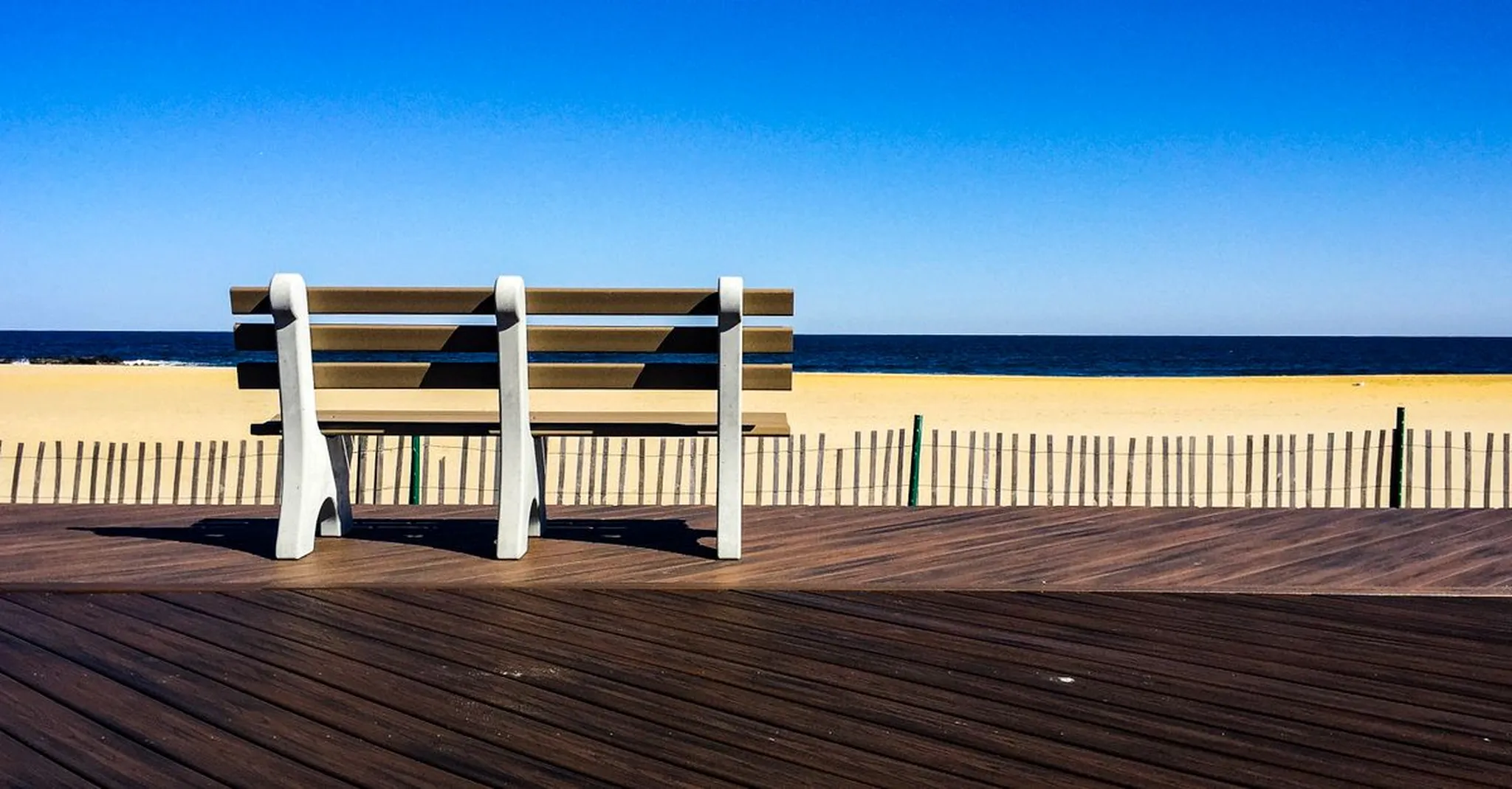 Empty wooden bench on a boardwalk overlooking a sandy beach and calm blue ocean under a clear sky