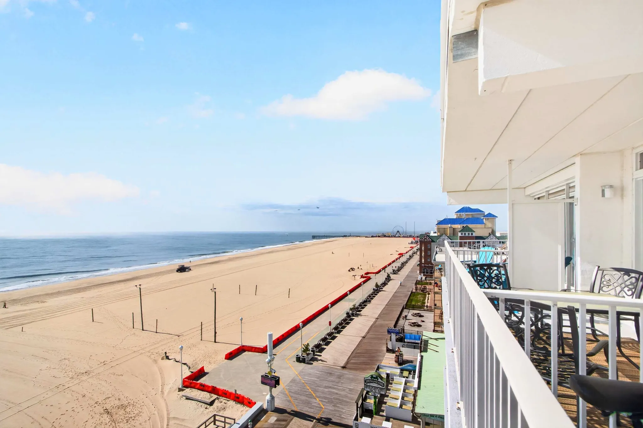 Balcony view overlooking a wide sandy beach, boardwalk, and ocean under a bright blue sky with wispy clouds