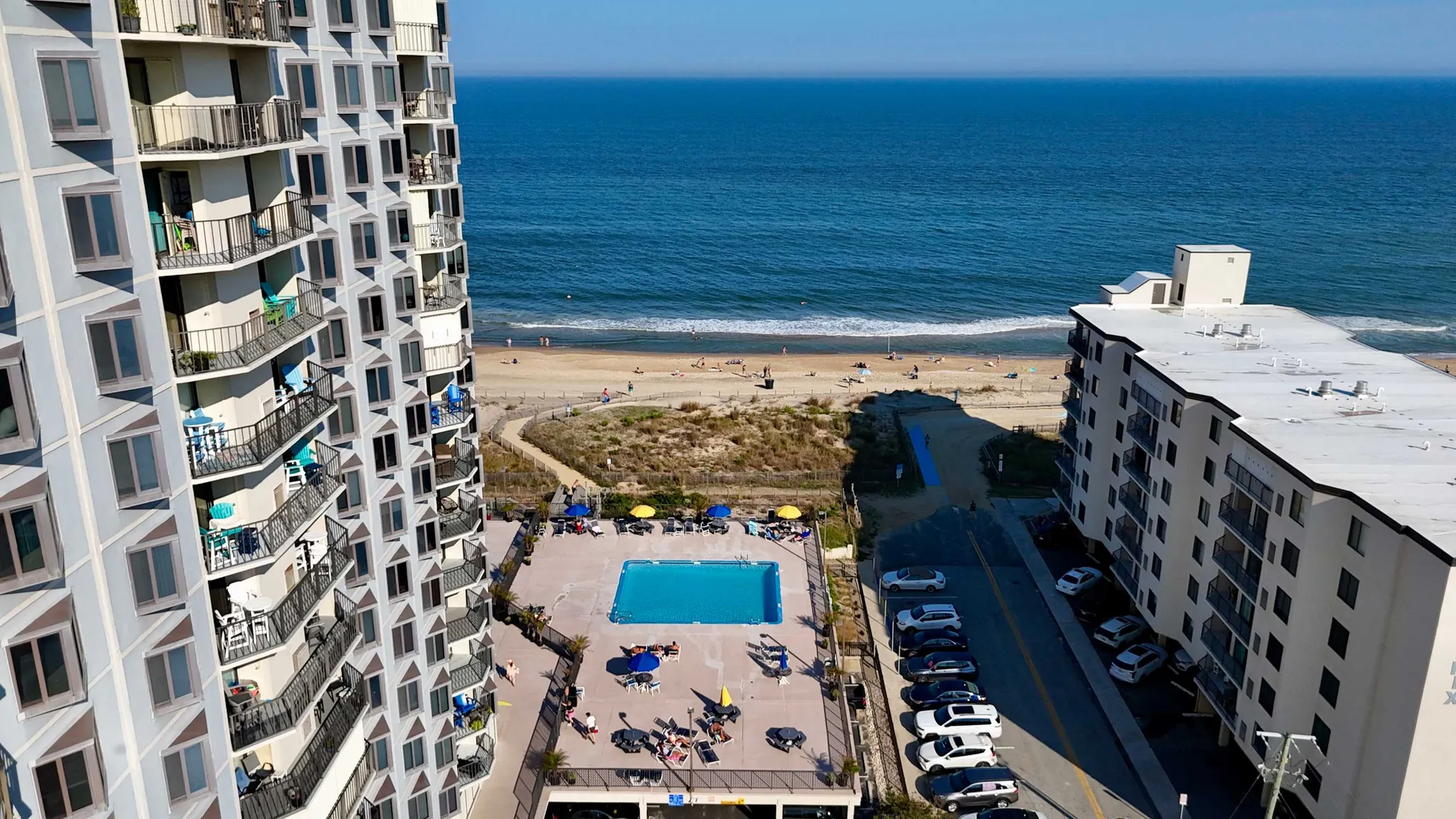Aerial view of beachfront condominium buildings with a rectangular pool, parking lot, and ocean shoreline under a clear blue sky