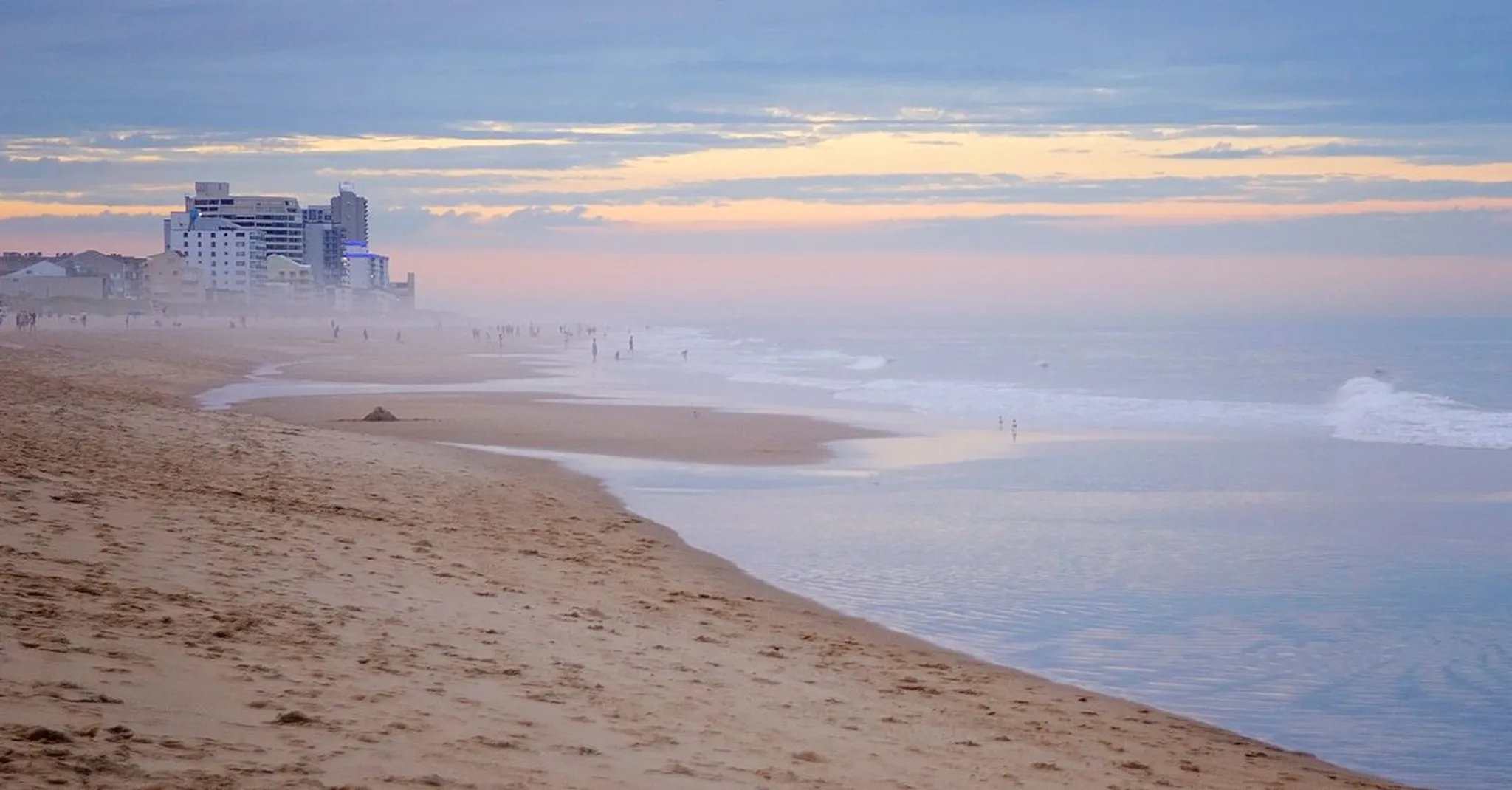 Wide sandy Atlantic beach at dusk with distant coastal high-rises, soft pastel sky, and gentle waves washing the shore