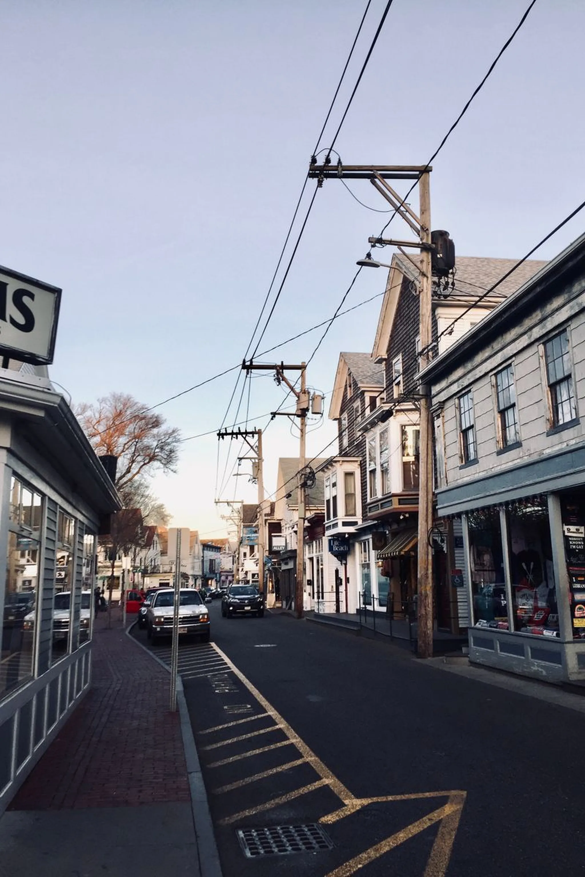 Quiet small-town main street at dusk with shops, parked cars, brick sidewalks and overhead power lines under a clear sky