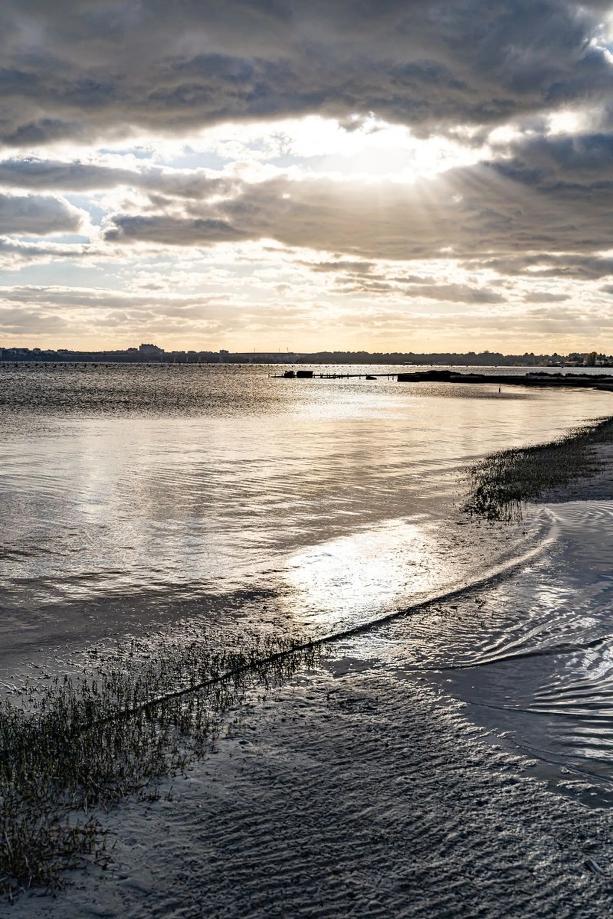 Sun breaking through clouds over calm bay with tidal flats, marsh grass, and distant shoreline at low tide