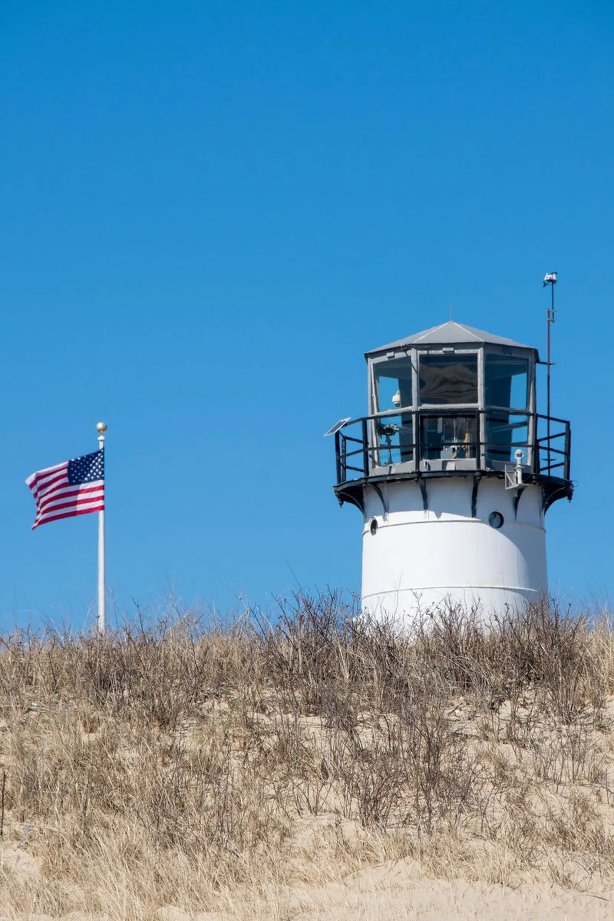 White lighthouse and American flag atop a grassy sand dune against a clear blue sky