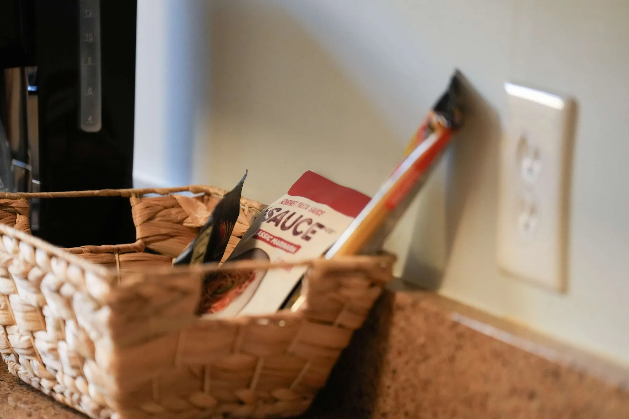 Woven basket of sauce packets and pantry items on a kitchen counter beside a coffee maker and wall outlet