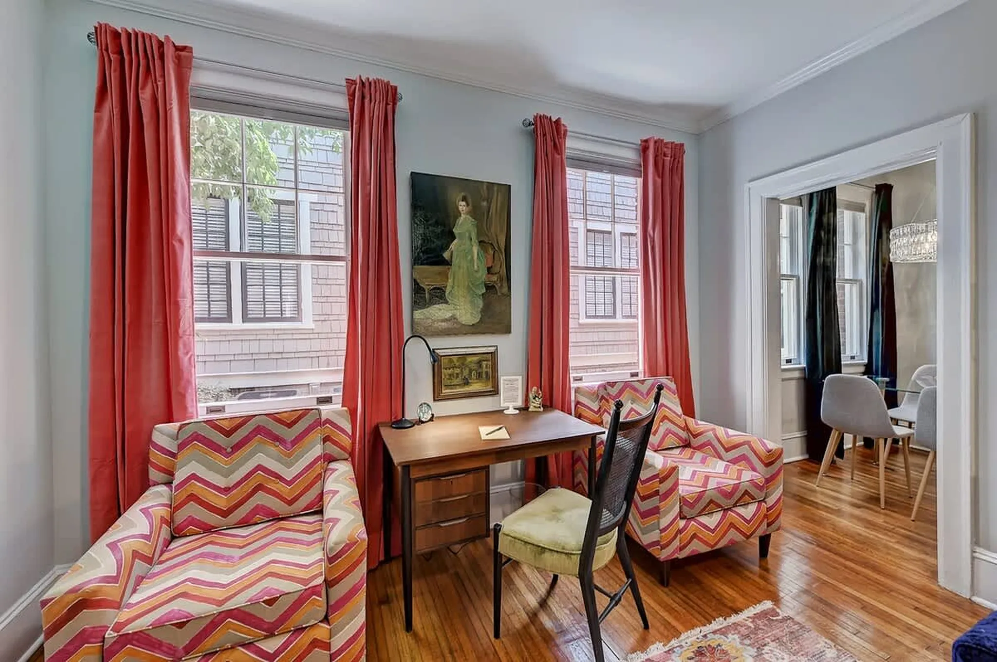 Sitting room with chevron-patterned armchairs, coral drapes, wooden desk, and framed portrait of a woman in white
