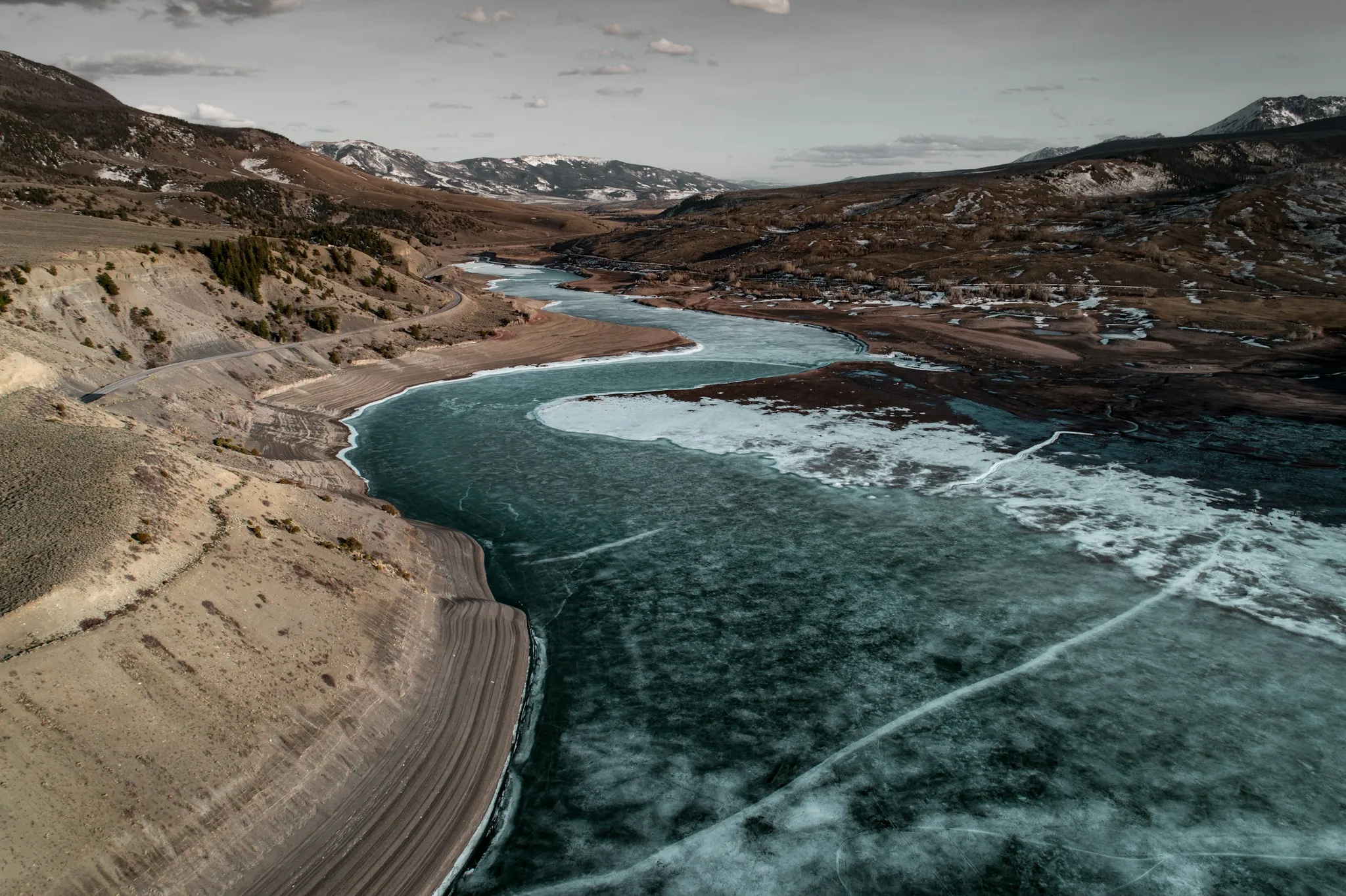Aerial view of a partially frozen river winding through barren mountain valley with distant town under a cloudy sky
