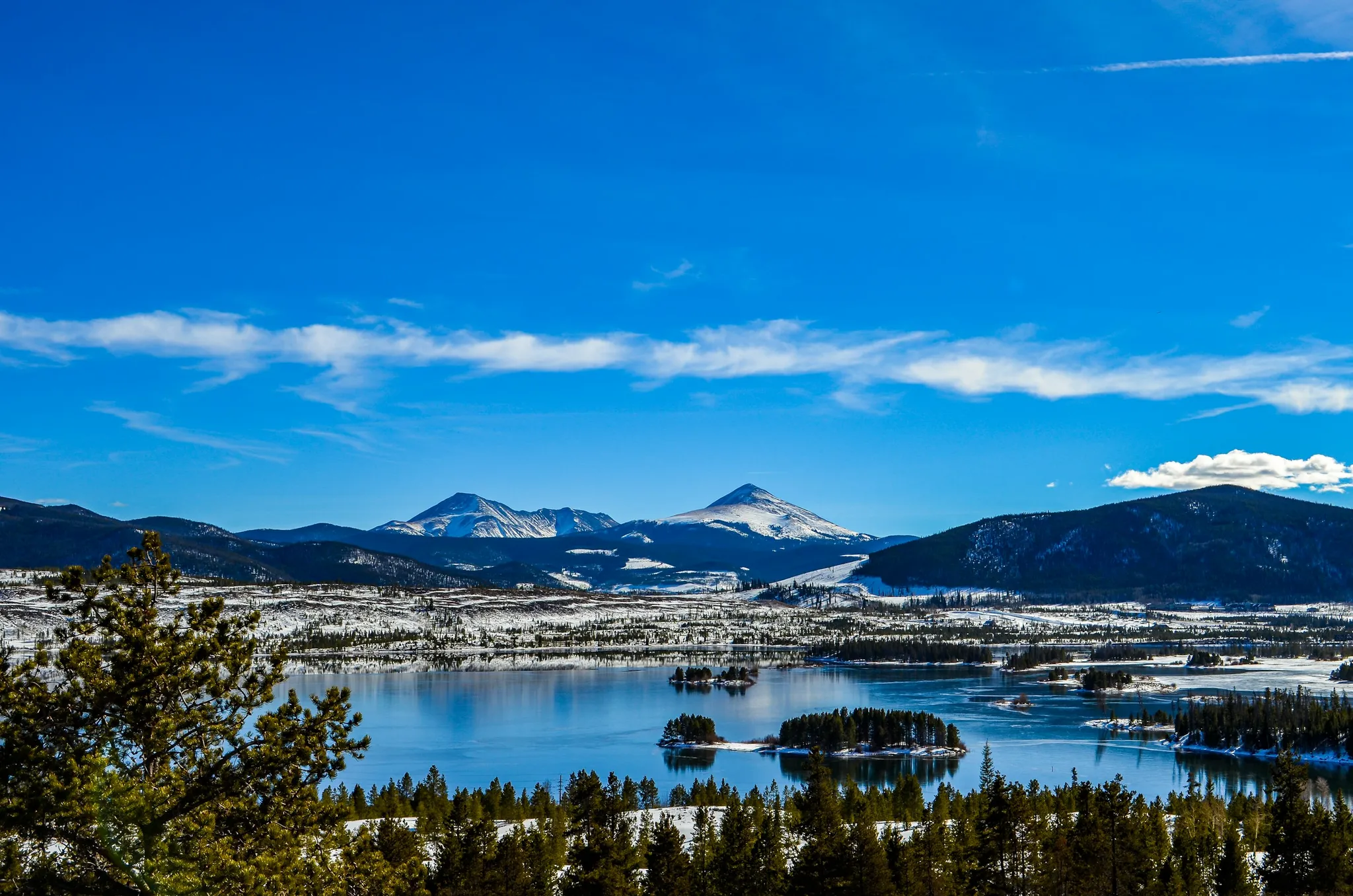 Snow-capped mountain peaks above a blue reservoir with pine trees and small islands under a clear sky