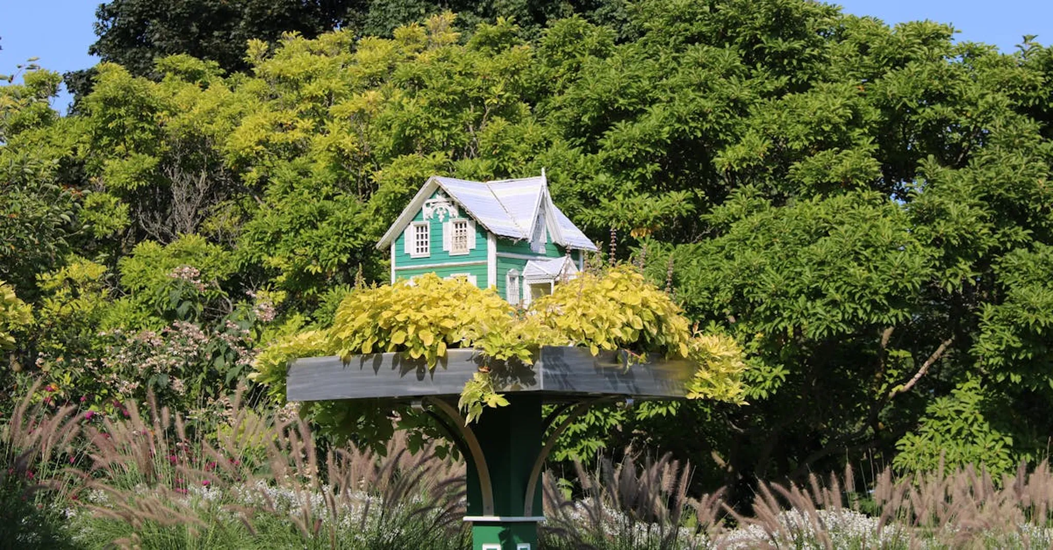 A quaint miniature green house sits atop a stand amidst verdant garden foliage.