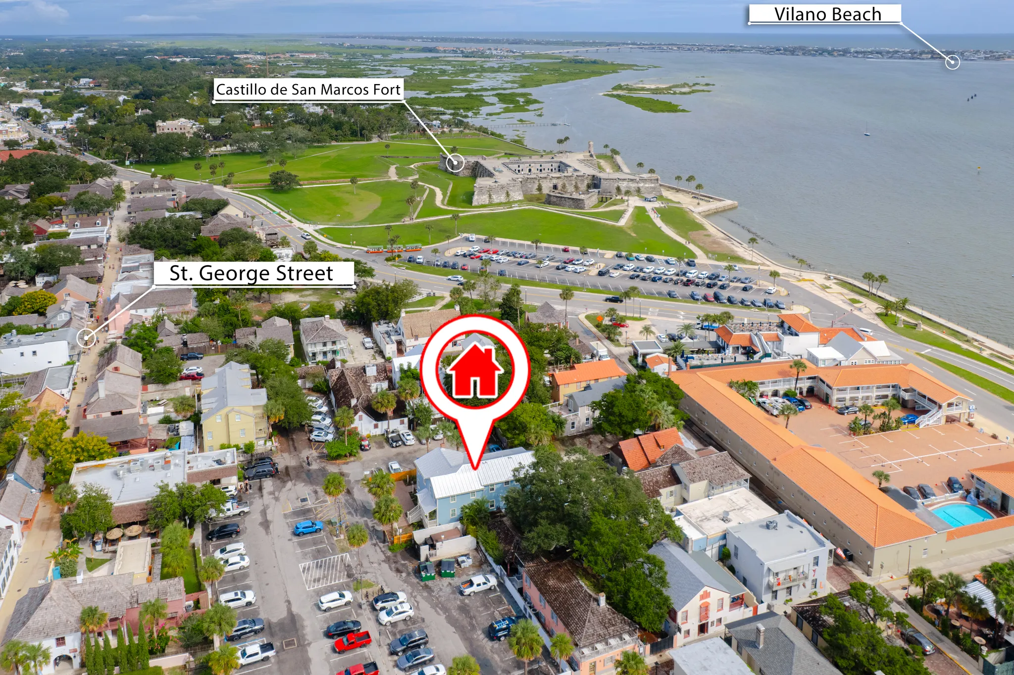 Aerial view of a coastal town with labeled landmarks, a waterfront fort, parking lots, and a red house pin marking a property location