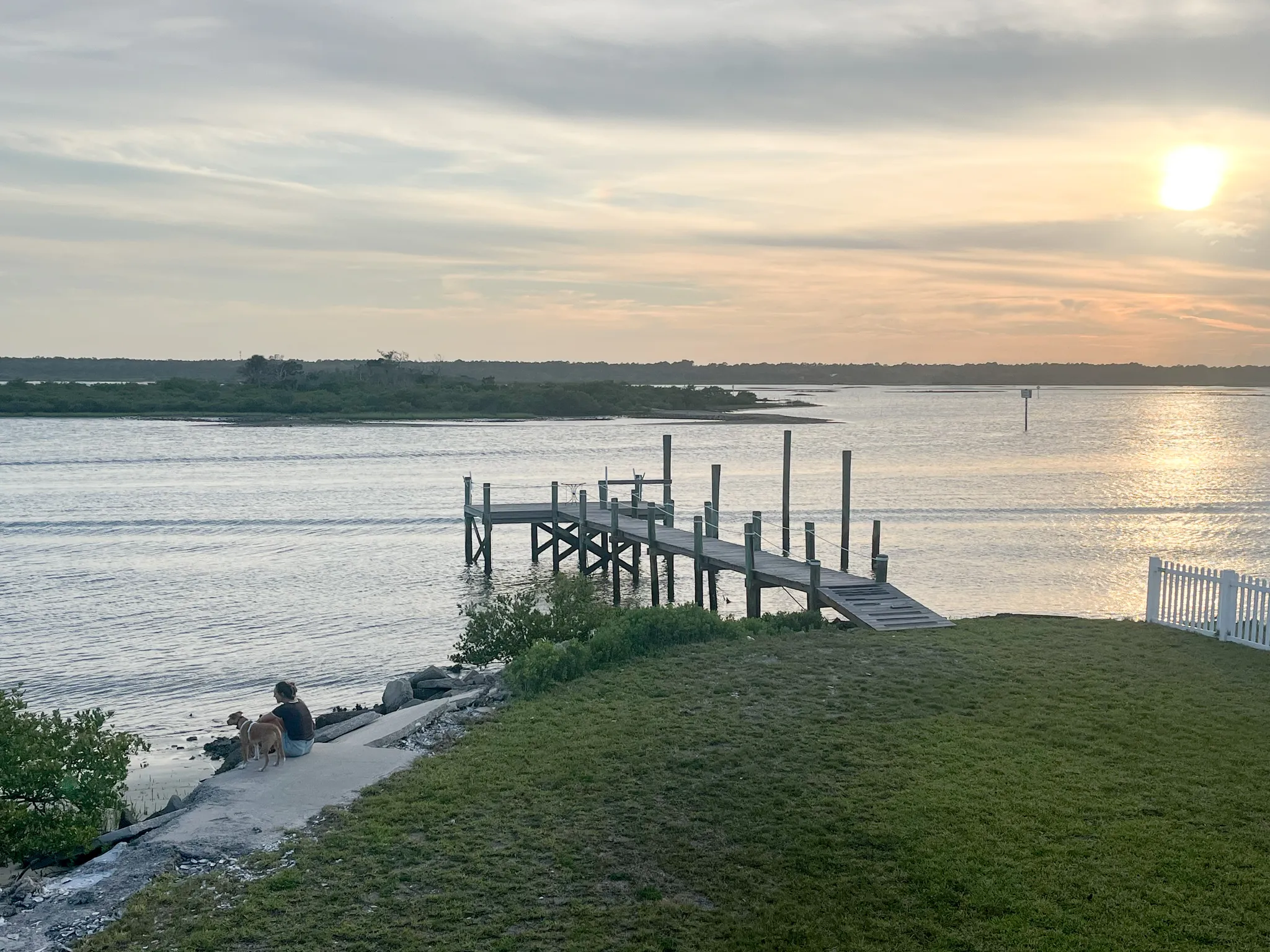 Person and dog sitting near a wooden dock at sunset, overlooking calm waters and distant tree-lined shoreline
