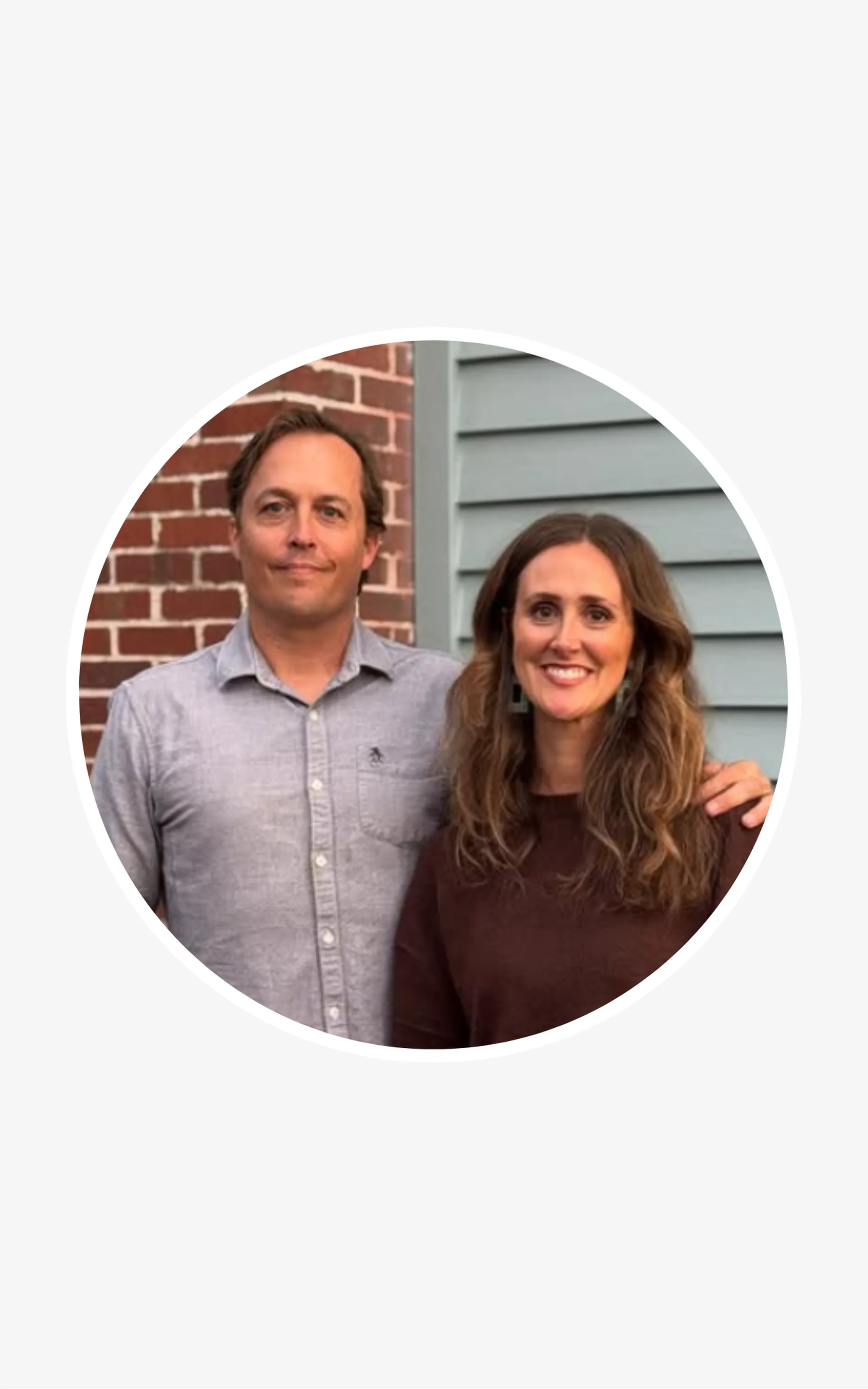 Smiling couple posing together outside a brick and gray-sided house, in a circular portrait frame