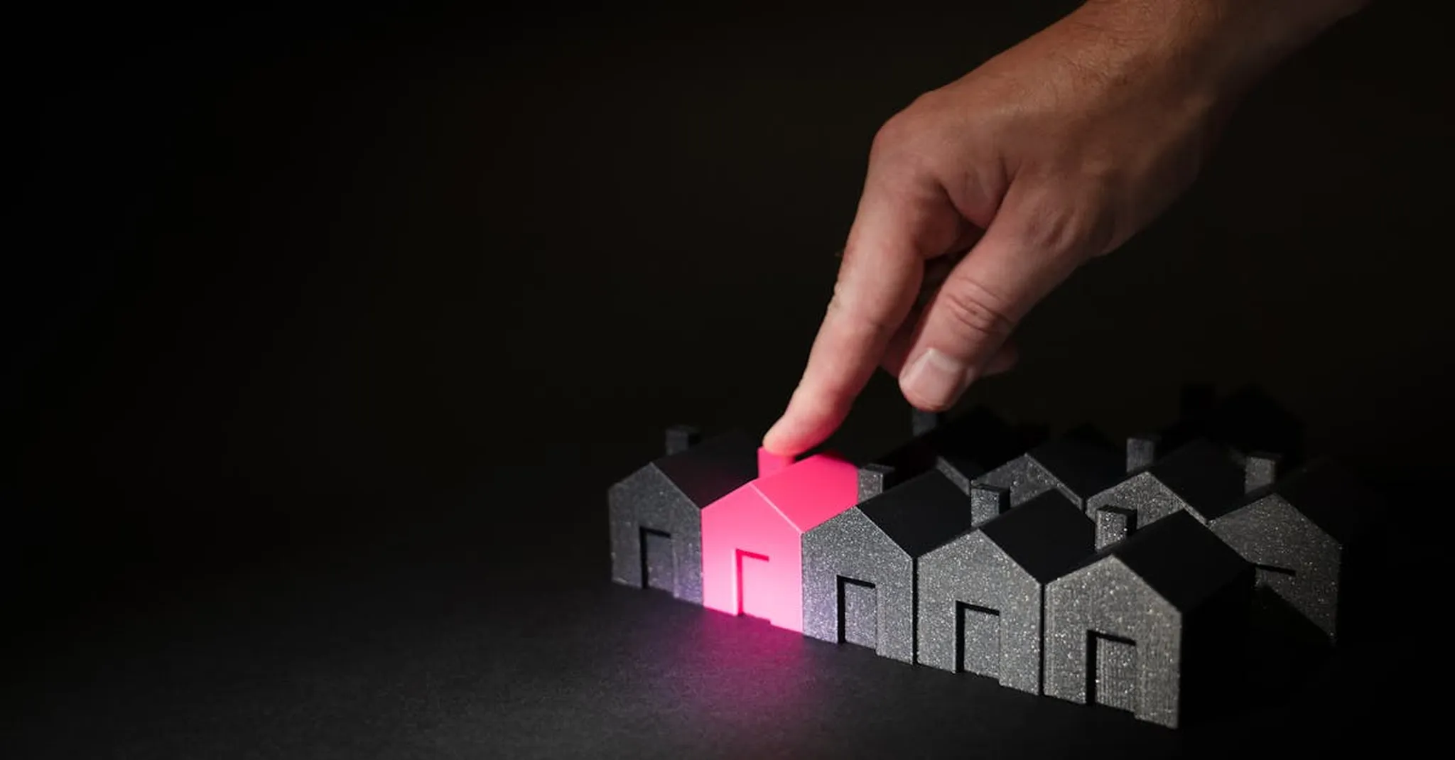 A hand selecting a glowing pink house among dark miniature houses, symbolizing real estate choices.